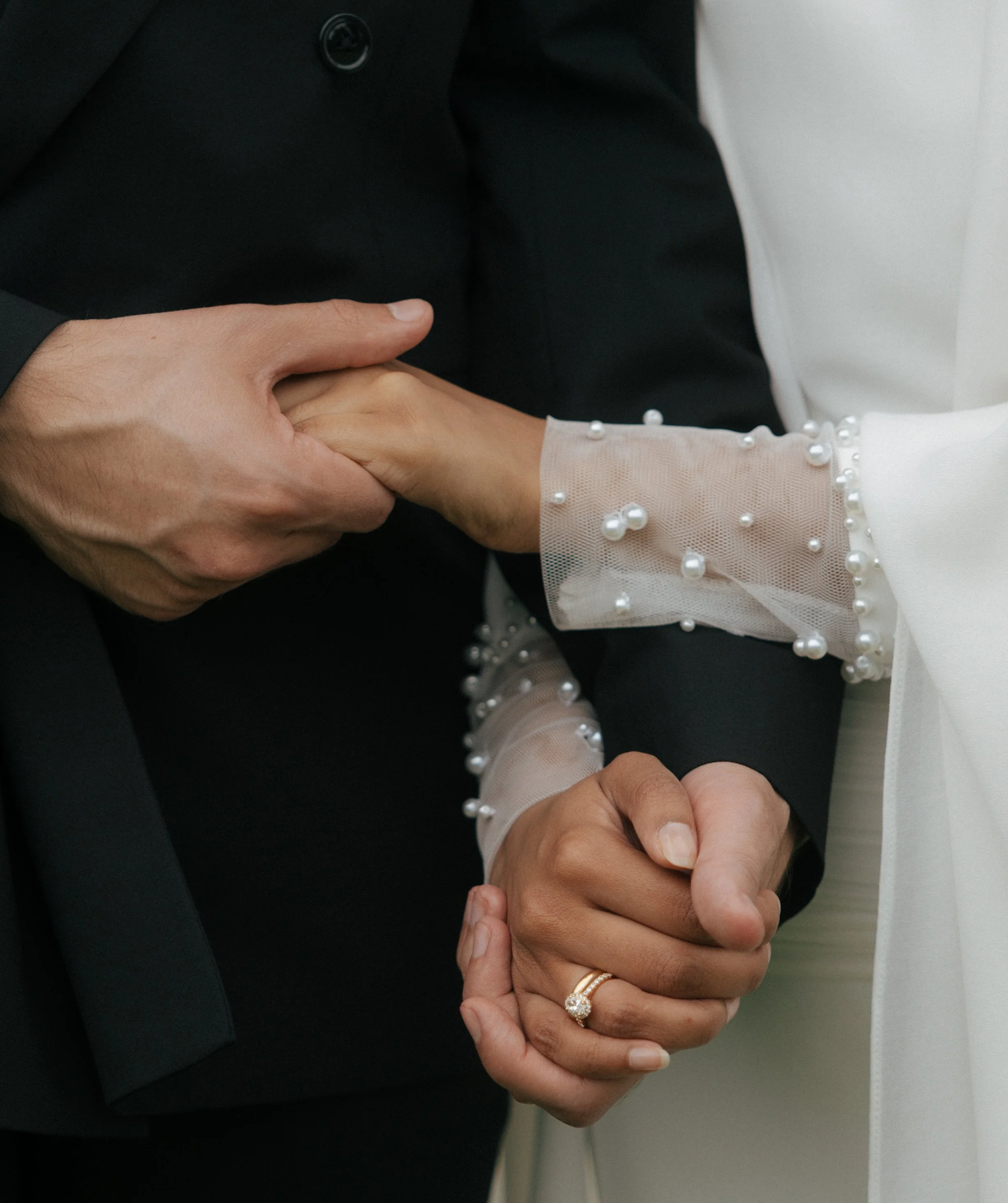 Close-up of a couple holding hands during a wedding, with the bride's hand showing a ring and her dress adorned with pearl embellishments.