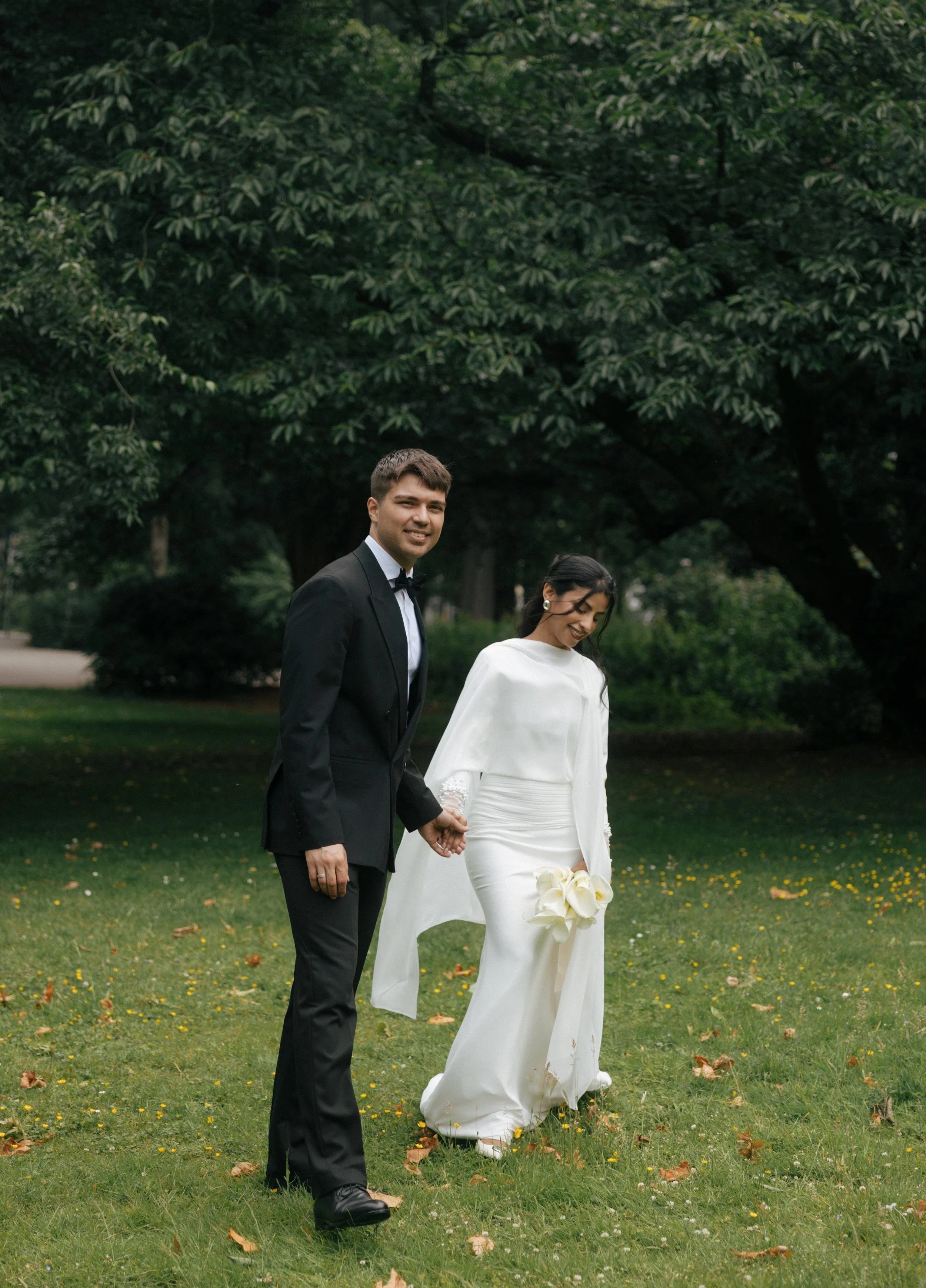 A bride and groom holding hands in a green park during their wedding, with trees in the background. The groom is wearing a black tuxedo, and the bride is in a white gown holding a bouquet of white calla lilies.