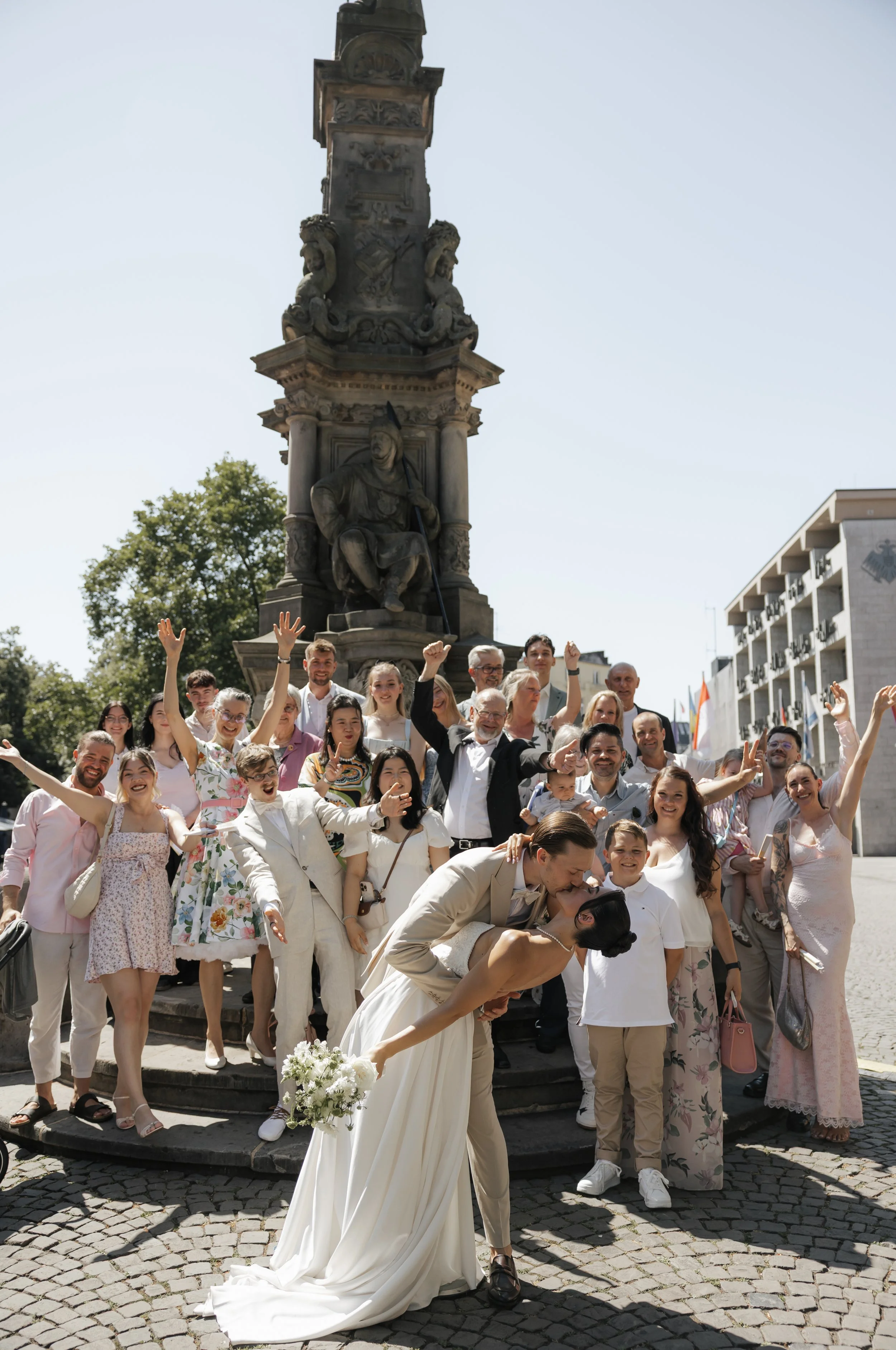 A wedding celebration with a bride and groom kissing in front of a crowd in an outdoor city square. The group is celebrating around a monument in the background on a sunny day.