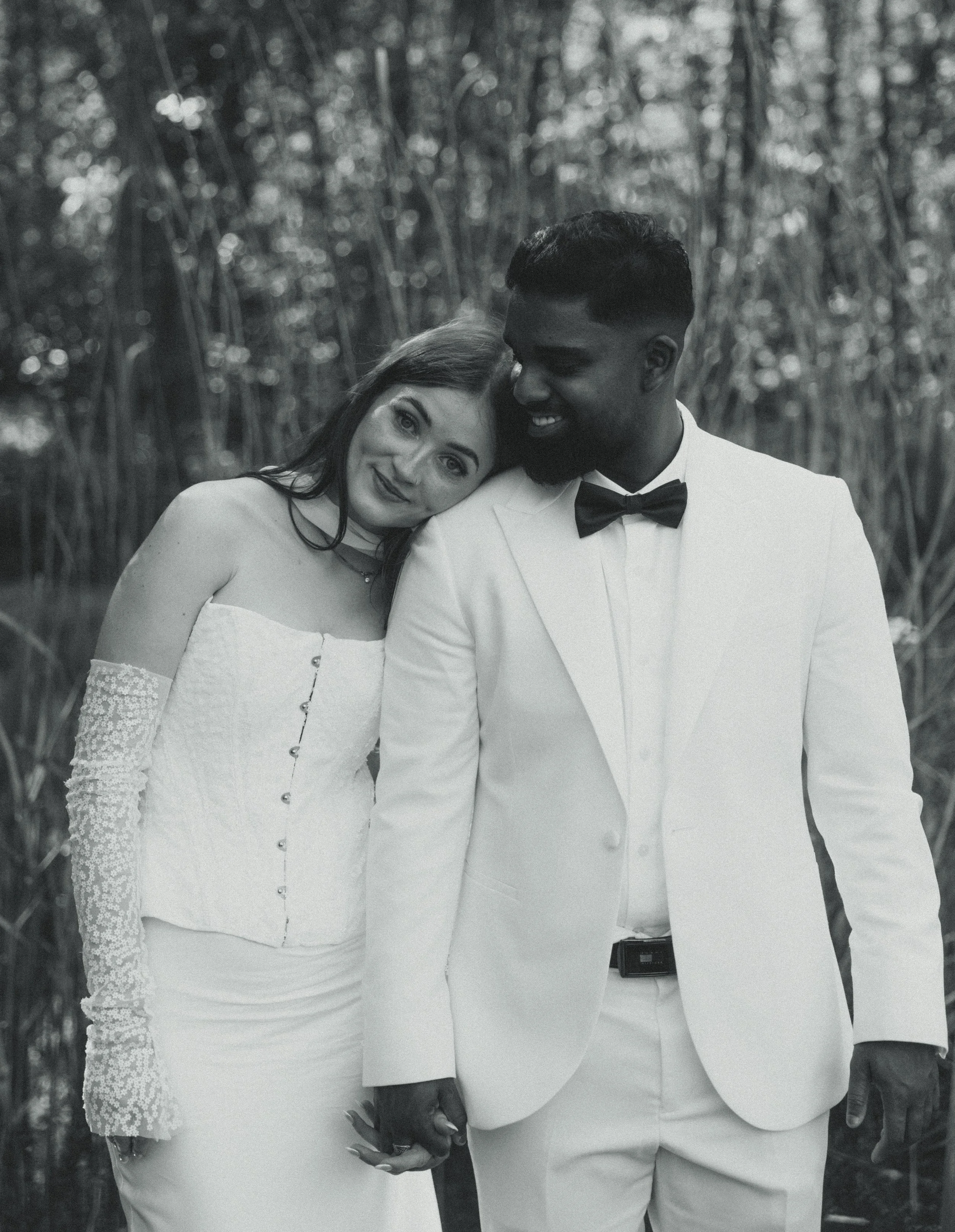 A black and white photo of a happy couple holding hands, standing close to each other outdoors amidst tall trees. The woman is dressed in an off-the-shoulder wedding gown with lace sleeves, and the man is wearing a white tuxedo with a black bow tie.