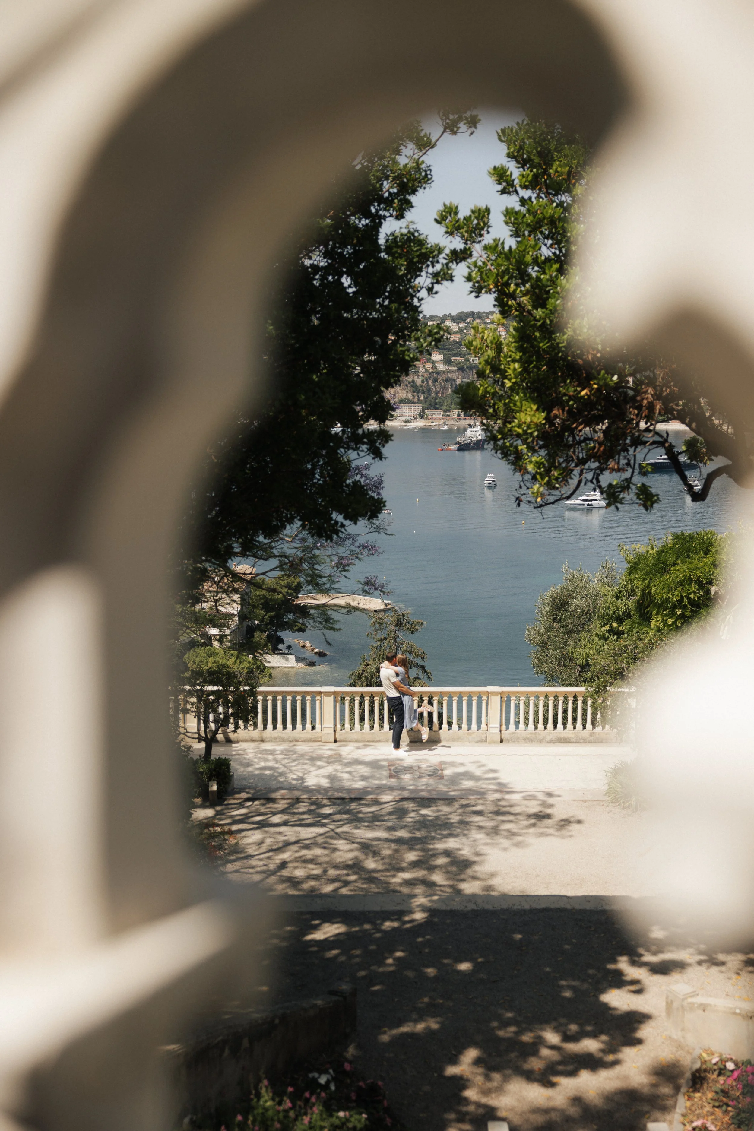 A couple embraces on a balcony overlooking a body of water with boats, framed by trees and a blurred foreground with a decorative railing.