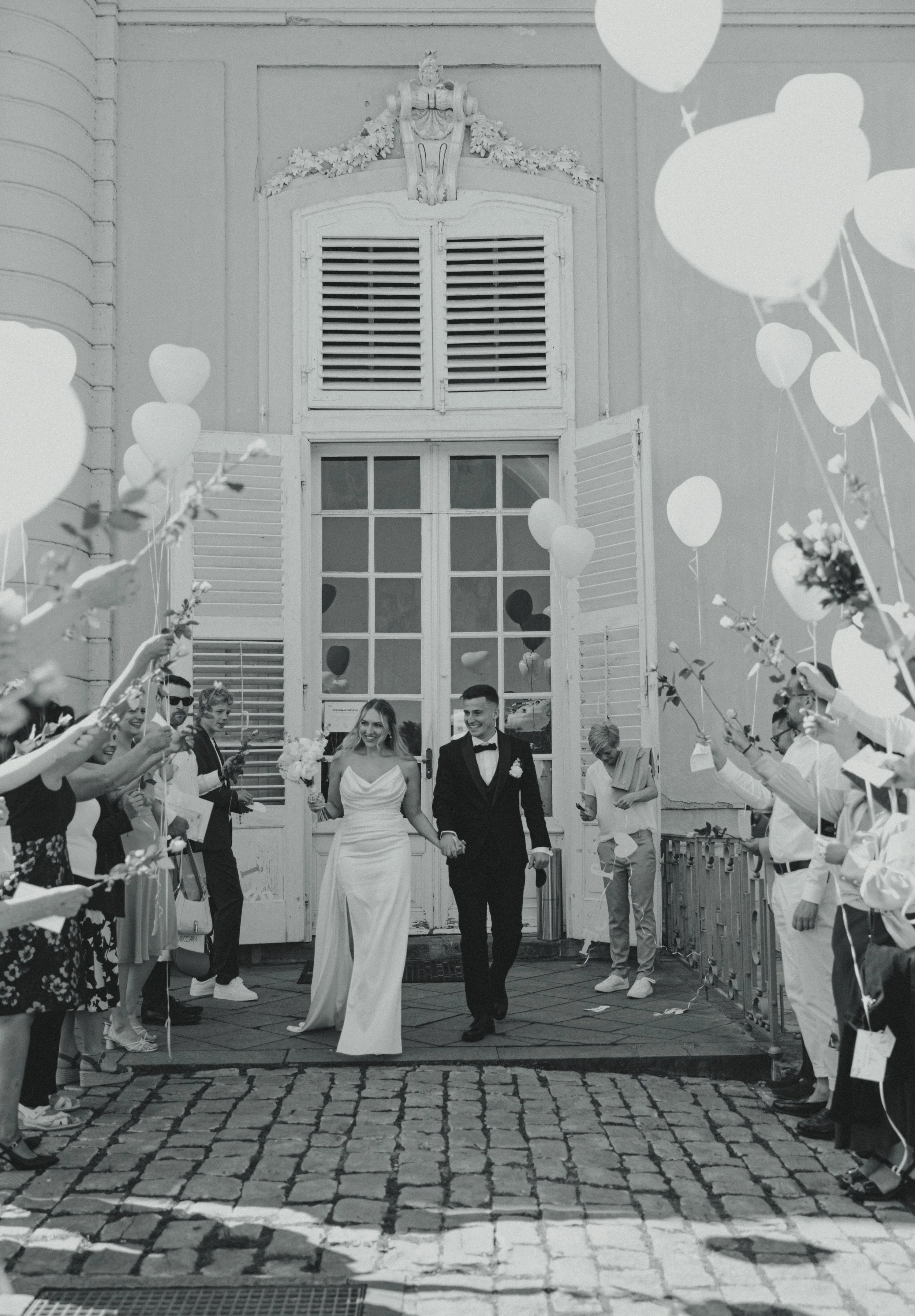 Black and white photo of a newlywed couple walking hand in hand through a aisle lined with guests holding balloons, outside a building with large windows and shutters.