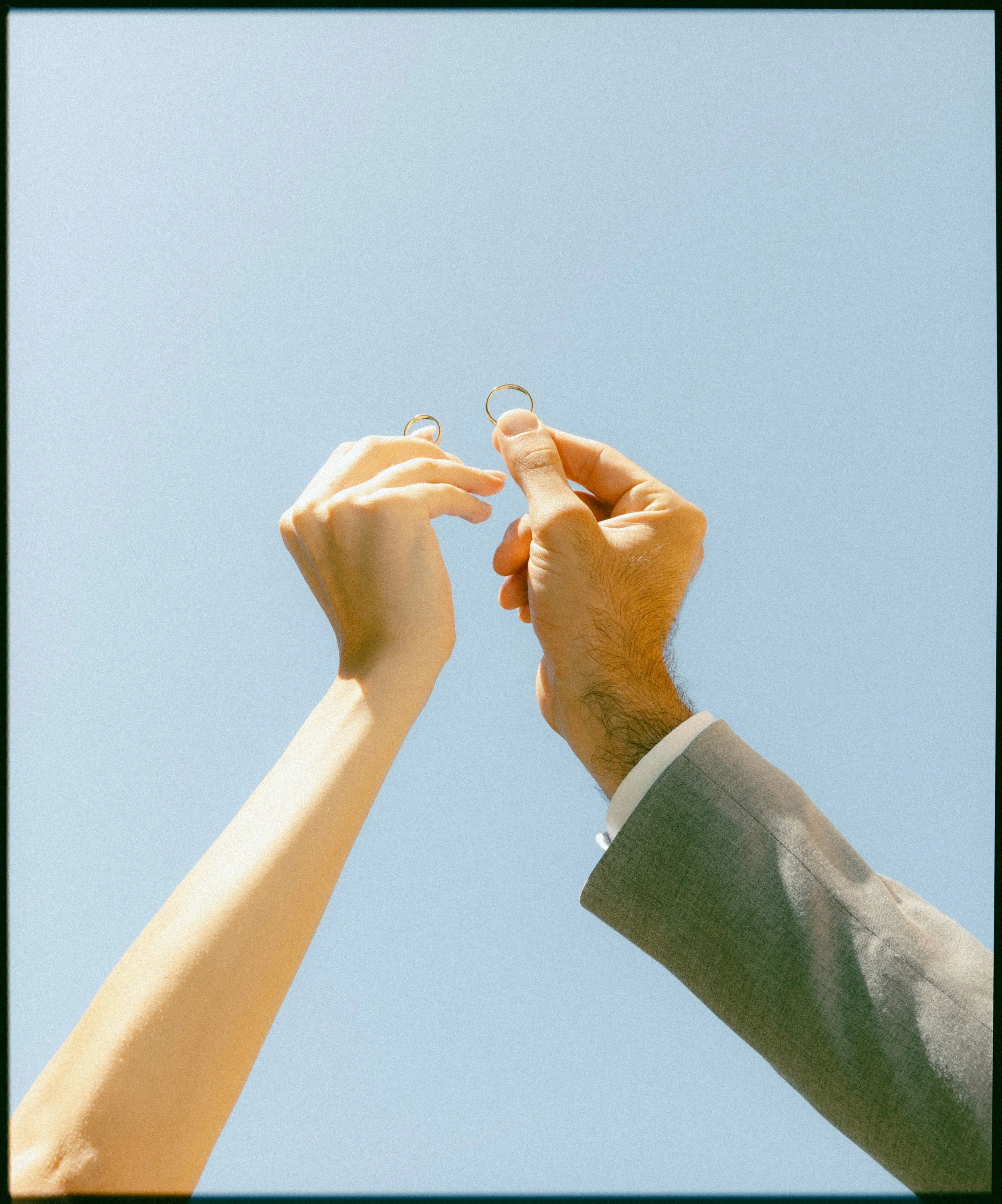Two hands, one female and one male in a suit, holding wedding rings against a clear blue sky.