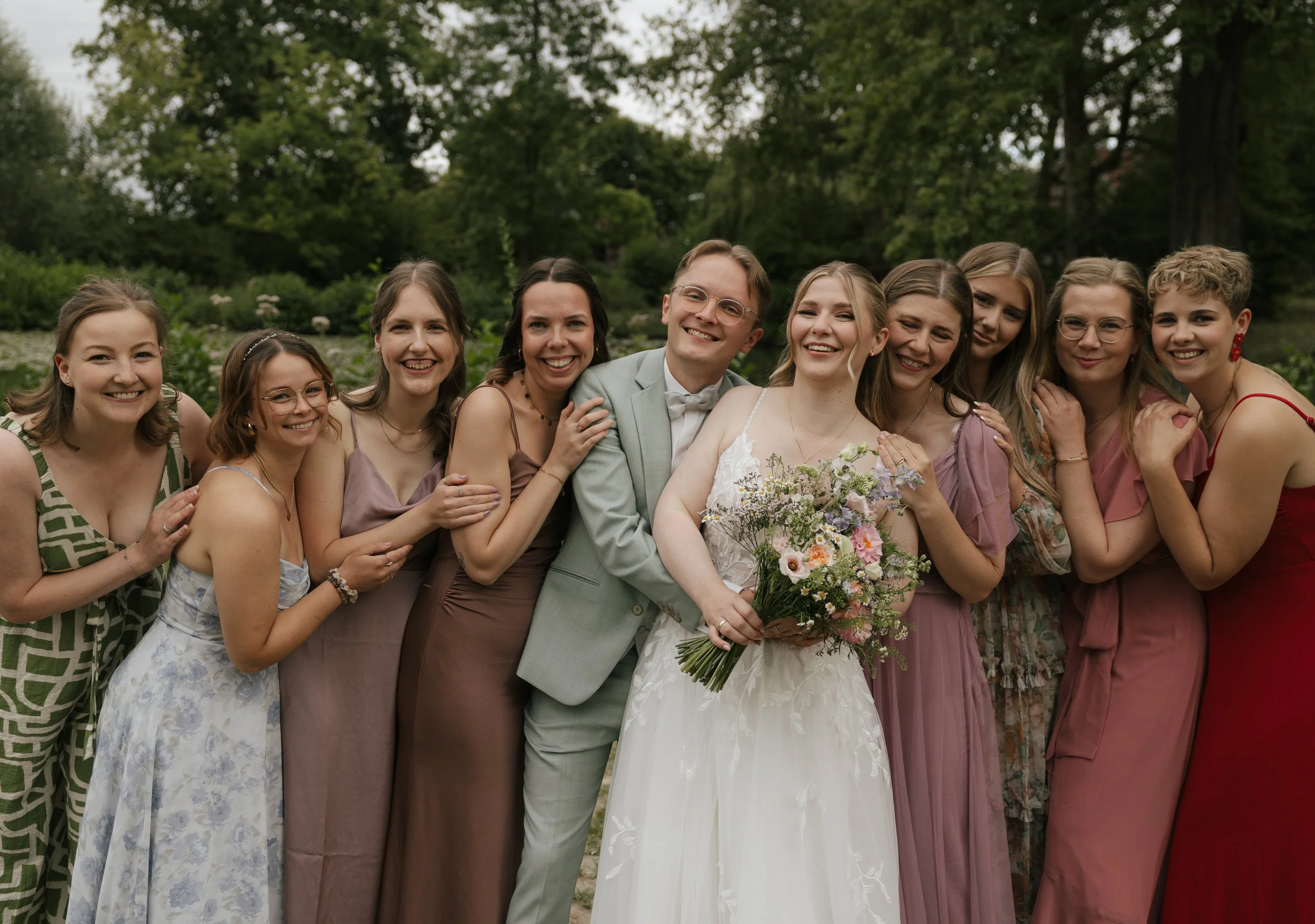 A group of eleven smiling people, including a bride in a white wedding dress holding a bouquet and a groom in a light gray suit, are posing outdoors with lush green trees. The group includes women dressed in various pastel and floral dresses.