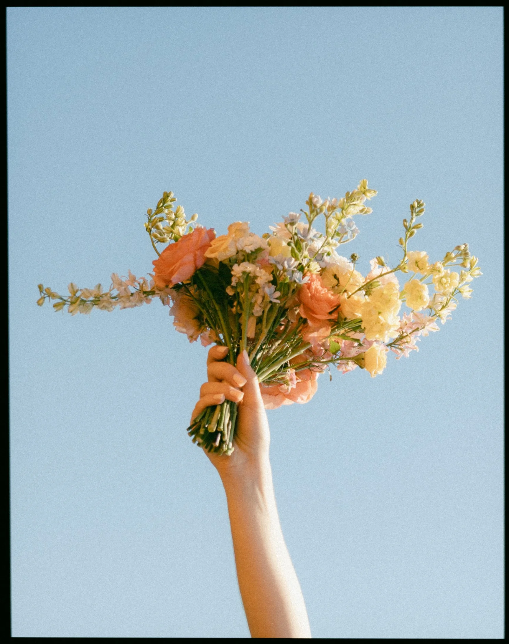 A hand holding a bouquet of mixed flowers against a clear blue sky.