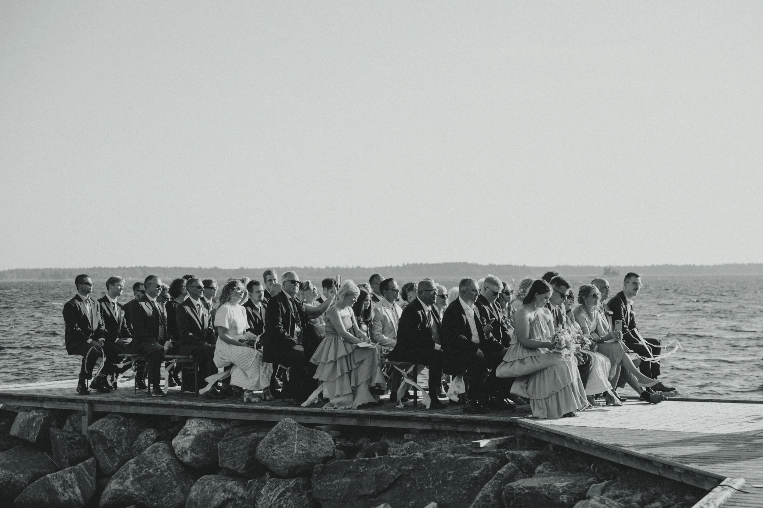 A group of people gathered on a dock by the water, dressed in formal attire, likely for a wedding ceremony.
