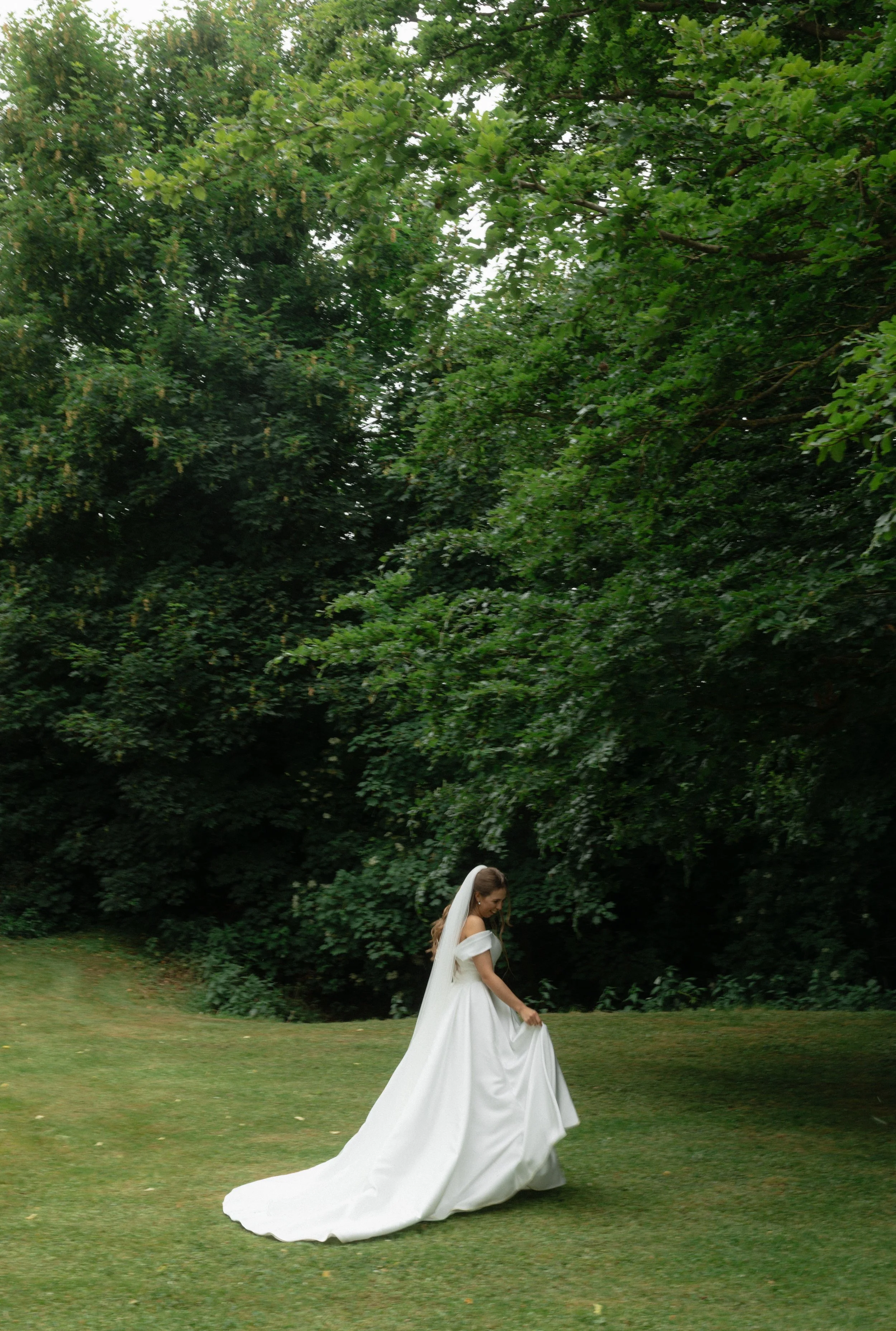 A bride in a white wedding dress with a long train and veil stands on grass near dense green trees, looking down as she lifts her dress slightly.