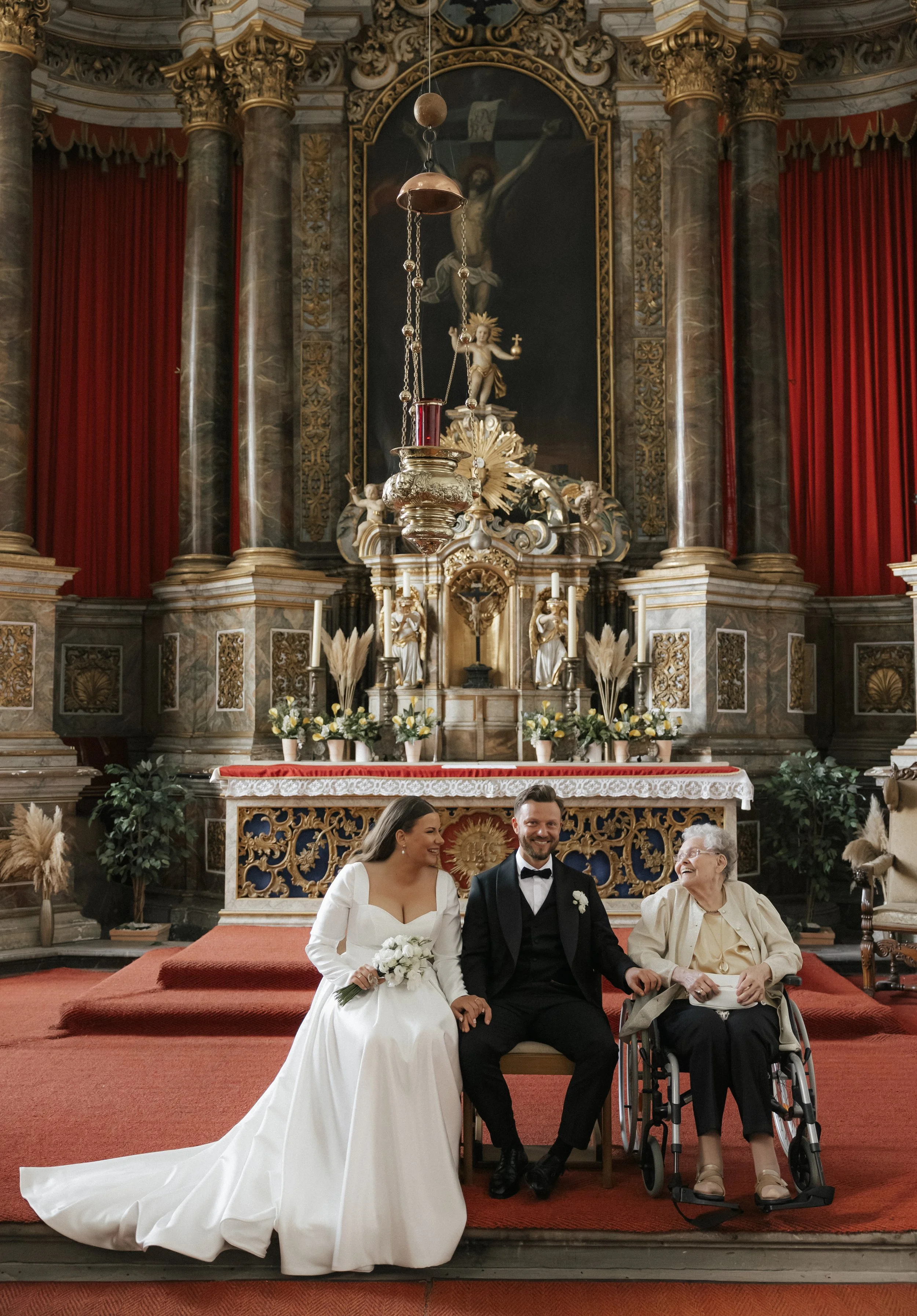 A wedding ceremony taking place inside a church, with a bride in a white gown, a groom in a black tuxedo, and an elderly woman in a wheelchair, sitting together on the church altar.