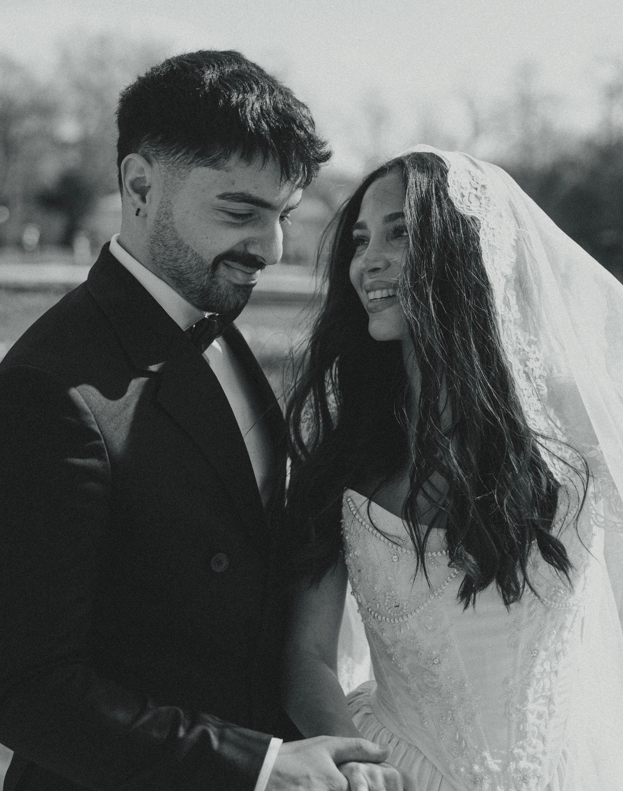 Black and white photo of a bride and groom standing outdoors, smiling and holding hands, with the bride wearing a veil and wedding dress. The groom is dressed in a tuxedo.