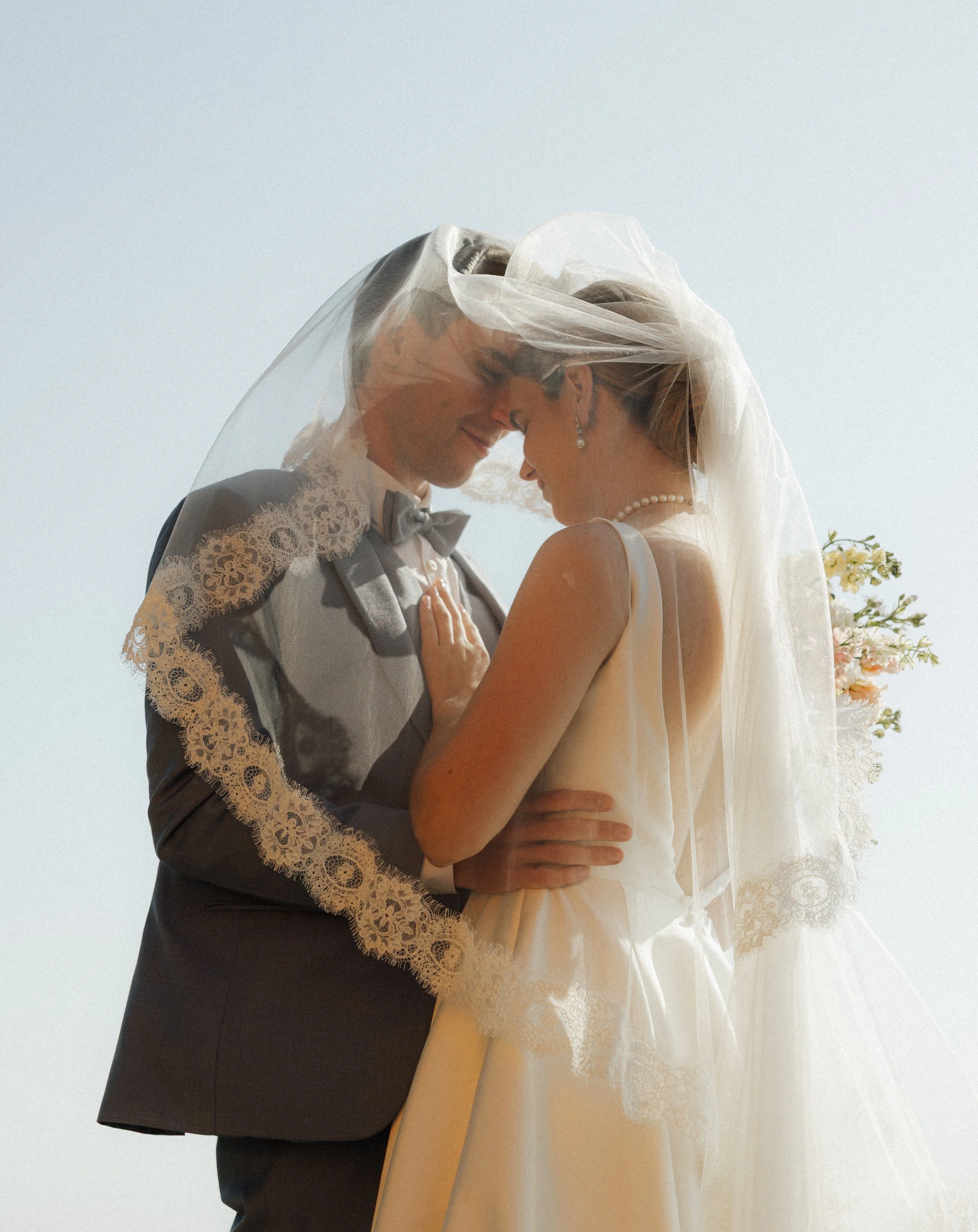 A bride and groom kiss under a veil on their wedding day, with a light blue sky background.