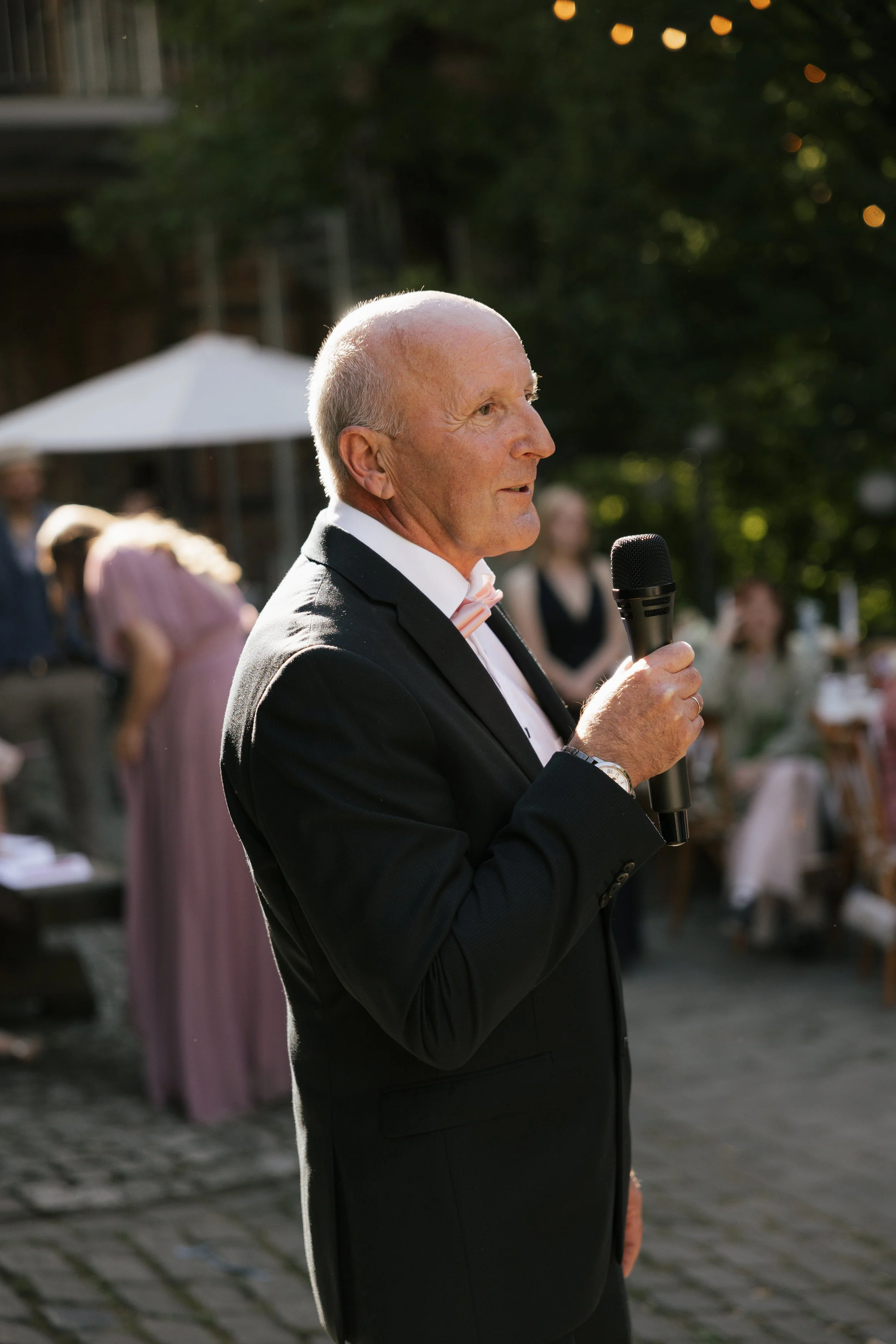 A senior man in a black suit and pink bow tie holding a microphone at an outdoor gathering during sunset.
