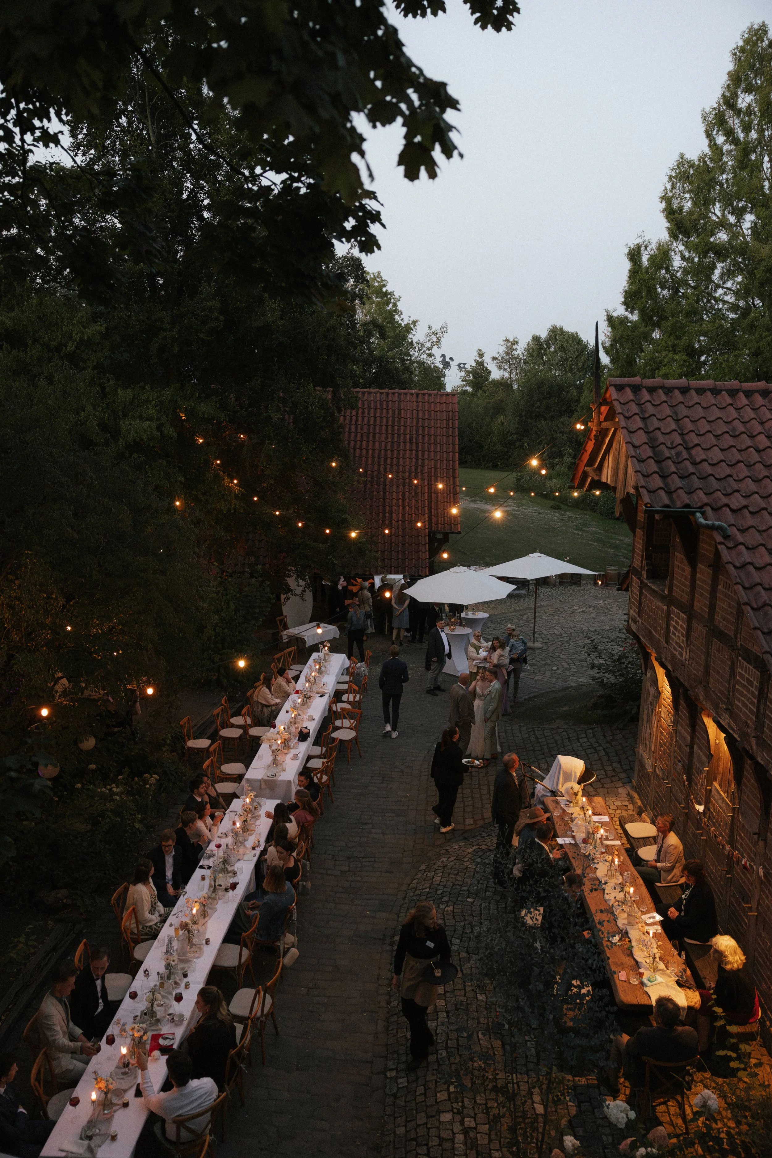 An outdoor evening gathering with long dining tables and string lights, surrounded by trees and rustic buildings.