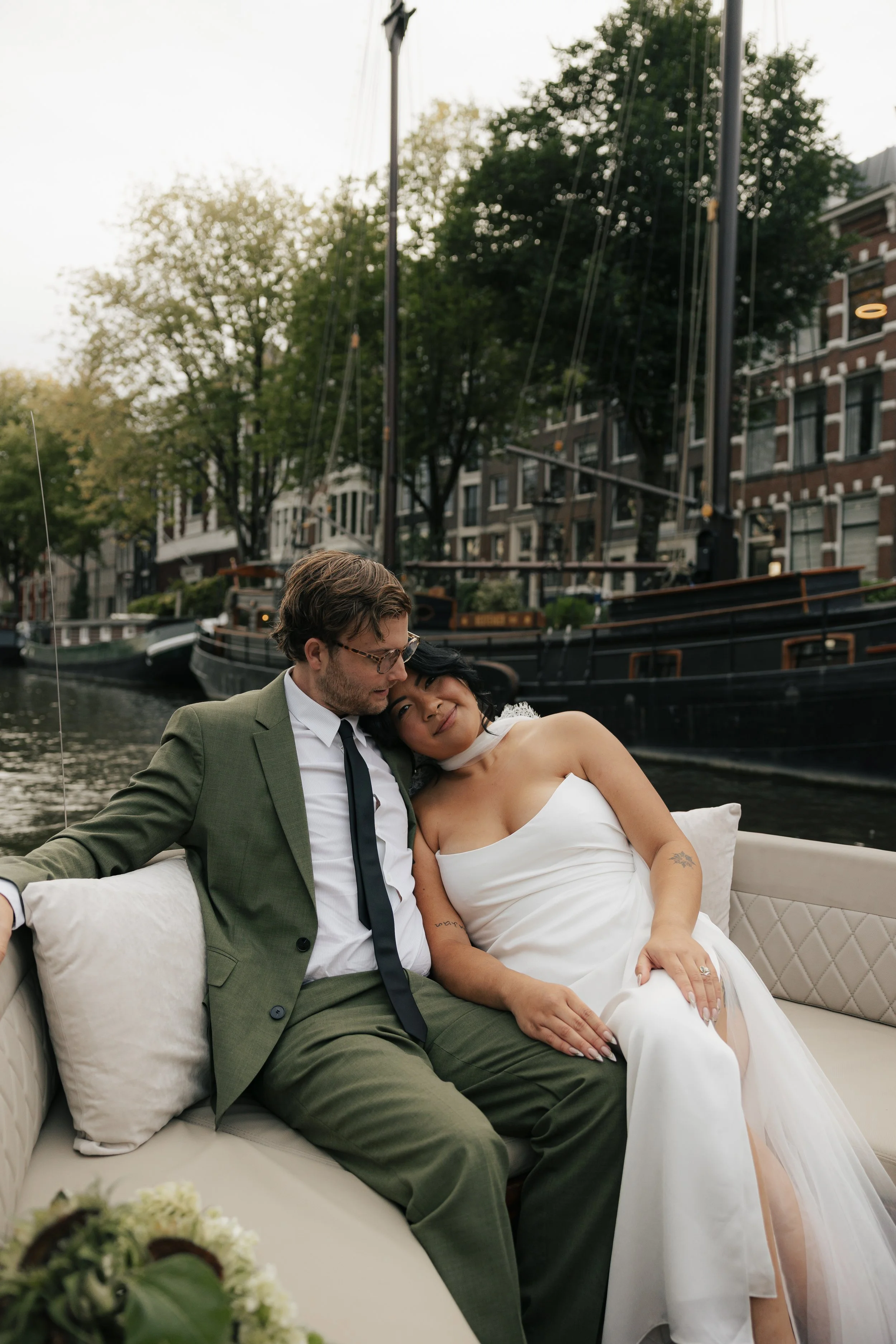 A couple dressed in wedding attire sitting on a boat with a canal and boats in the background.
