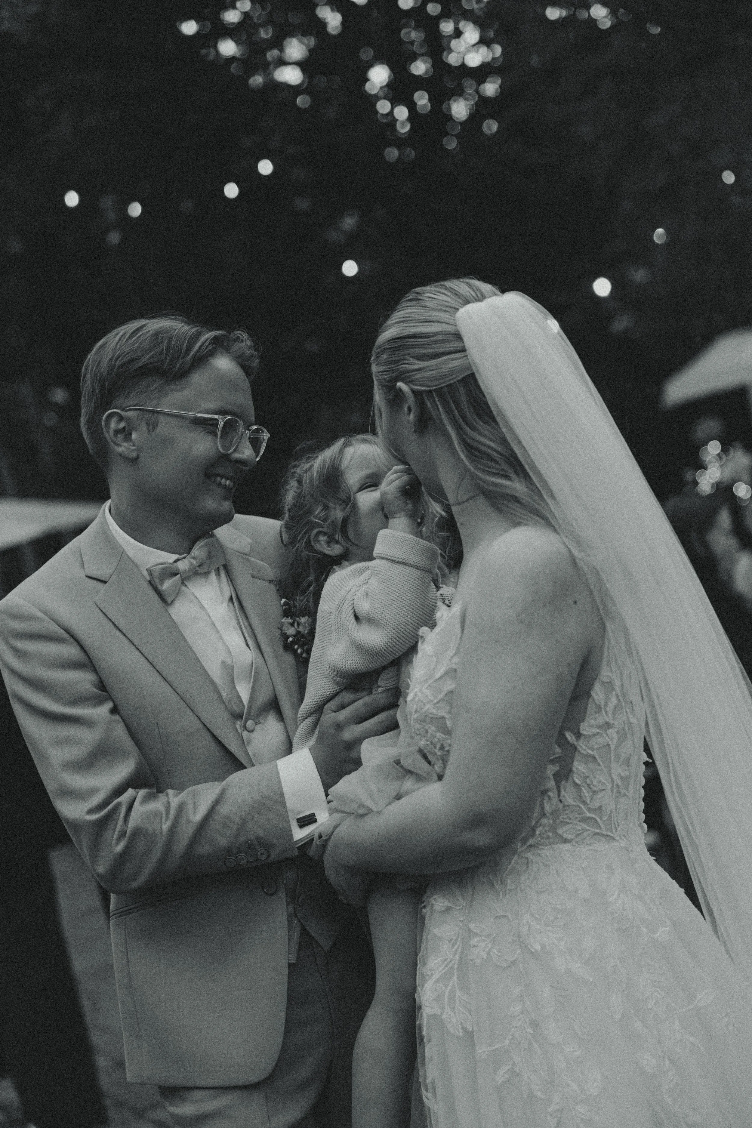A black and white photo of a wedding scene showing a couple, a bride in a lace wedding dress and a groom in a suit, holding a young girl. The girl is touching the bride's face and smiling, and all are smiling. The background has out-of-focus lights.