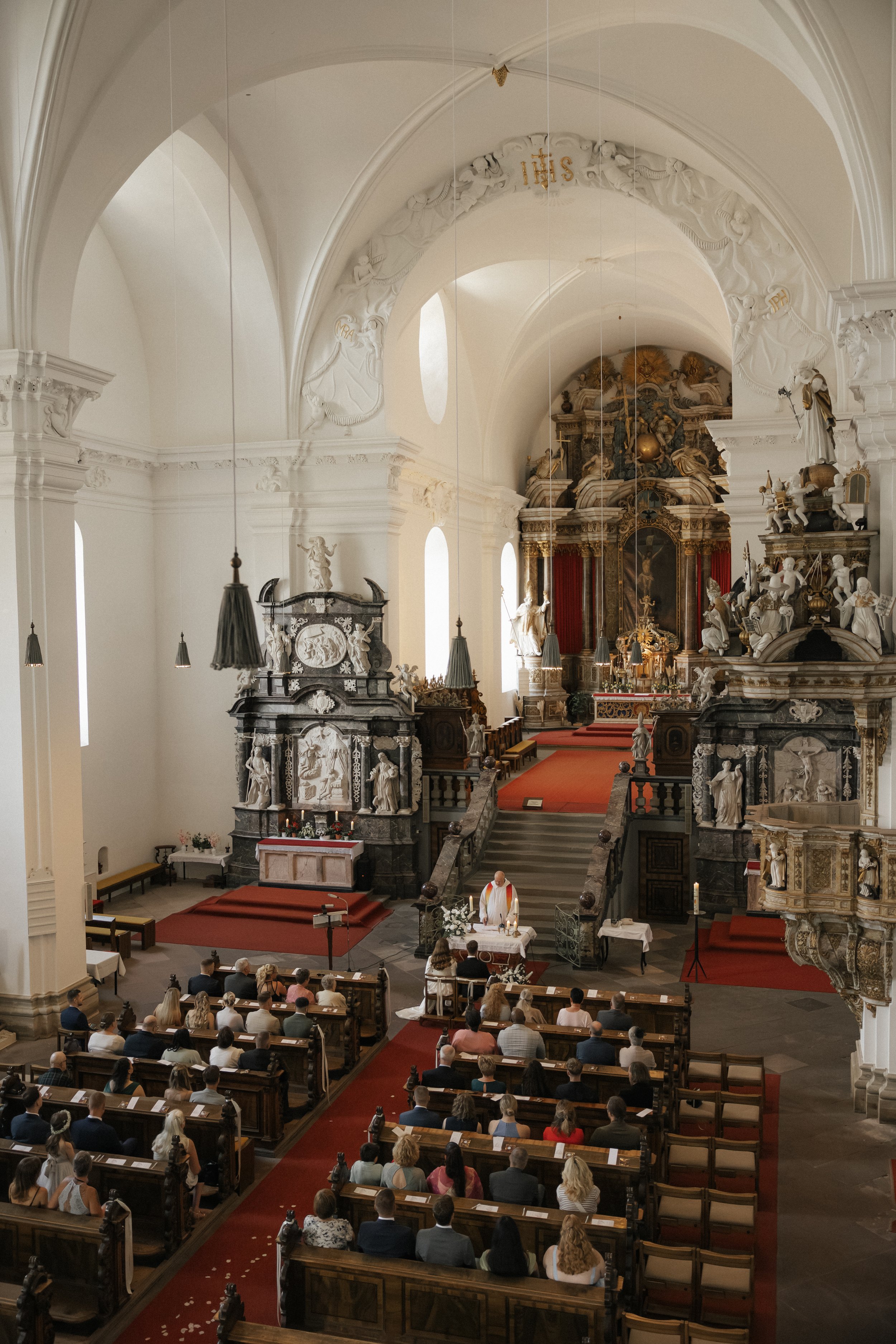 Interior of a church with a congregation seated in pews facing the altar where a priest is conducting a service. The church is decorated with intricate white and gold sculptures and decorations, with high arched ceilings and large windows letting in natural light.