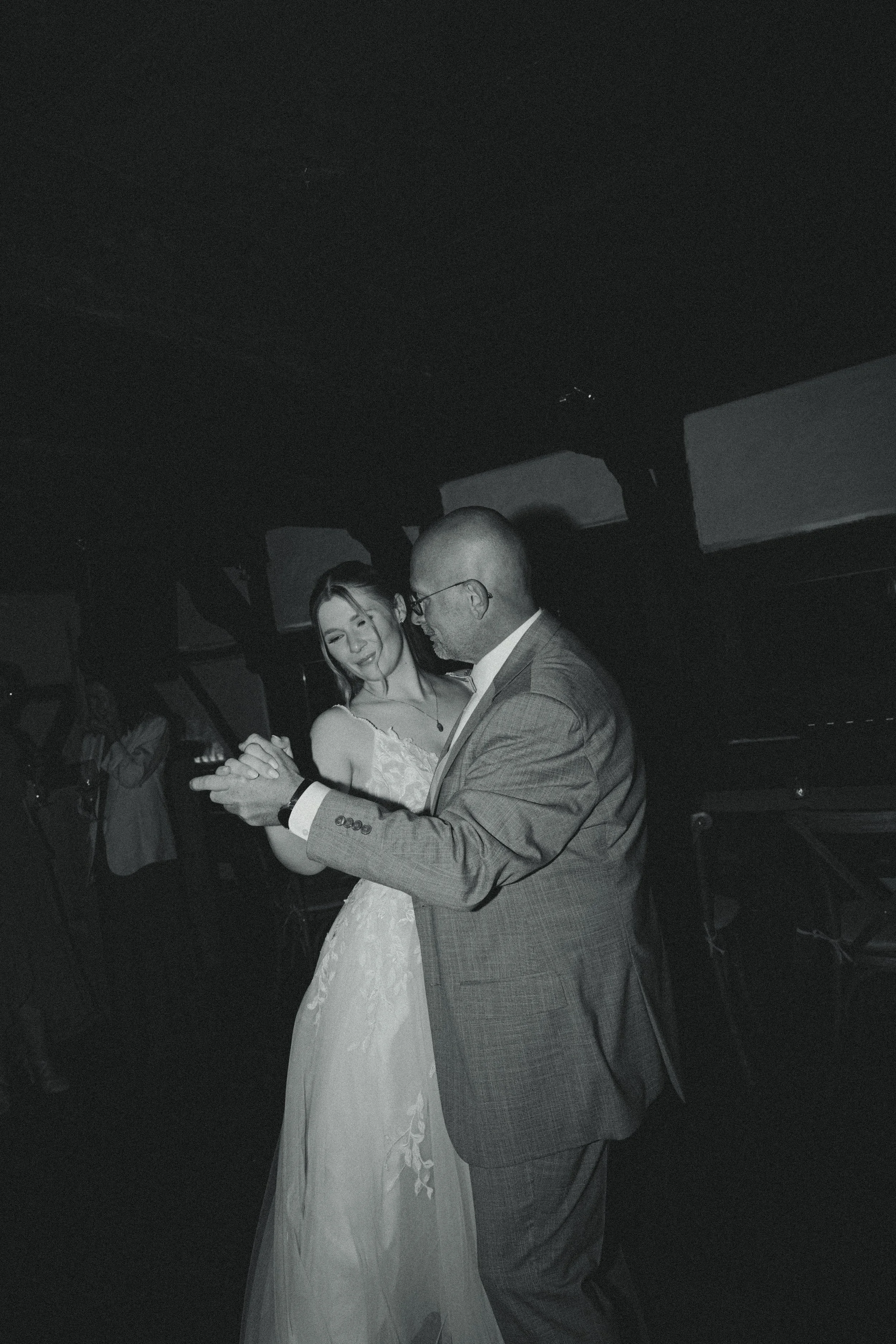 A black and white photo of a bride in a wedding dress dancing with a groom in a suit at a wedding reception.