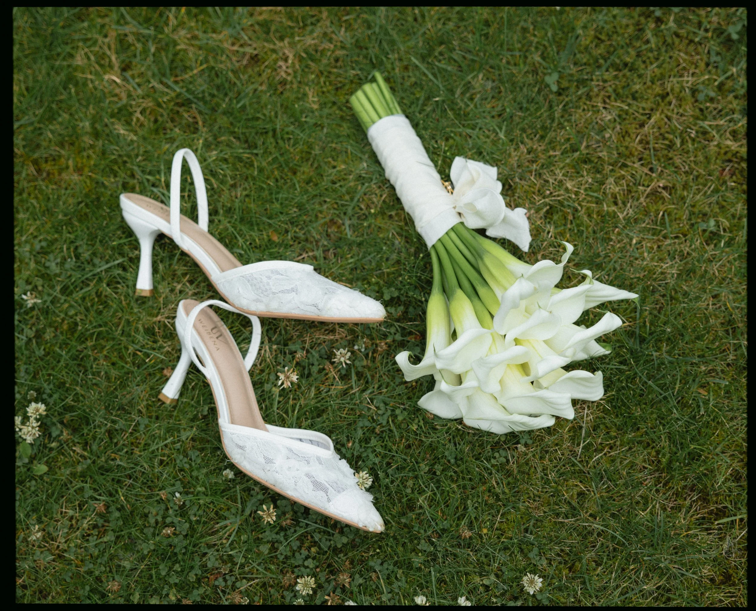 A pair of white lace high heels and a bouquet of white calla lilies on green grass.