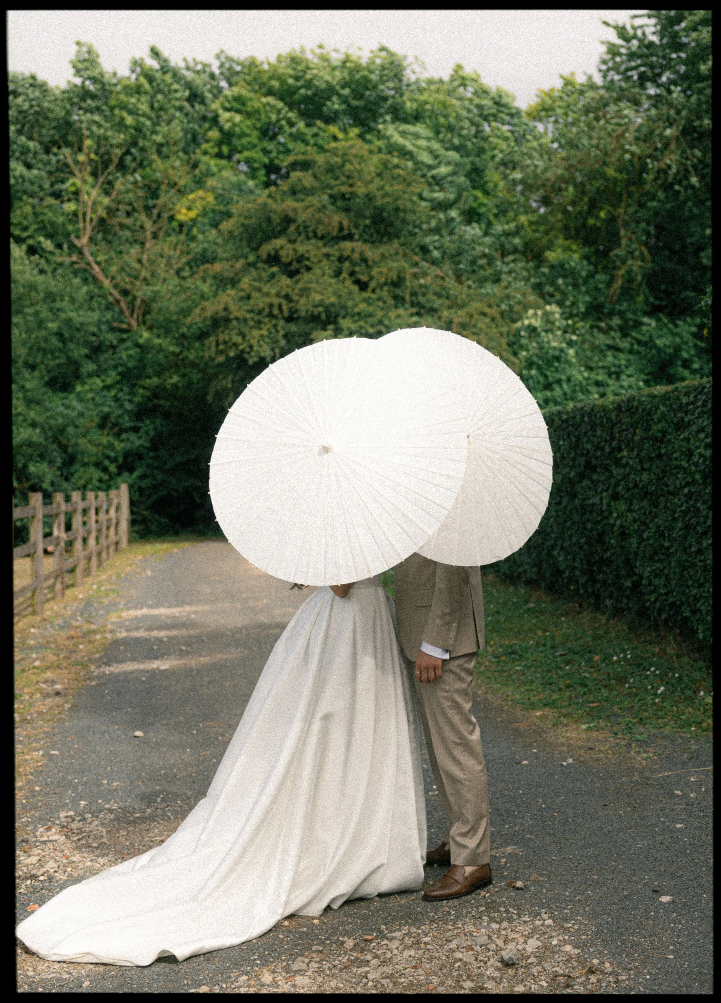 A bride and groom standing close together on a gravel path, holding white parasols over their faces, with trees and greenery in the background.