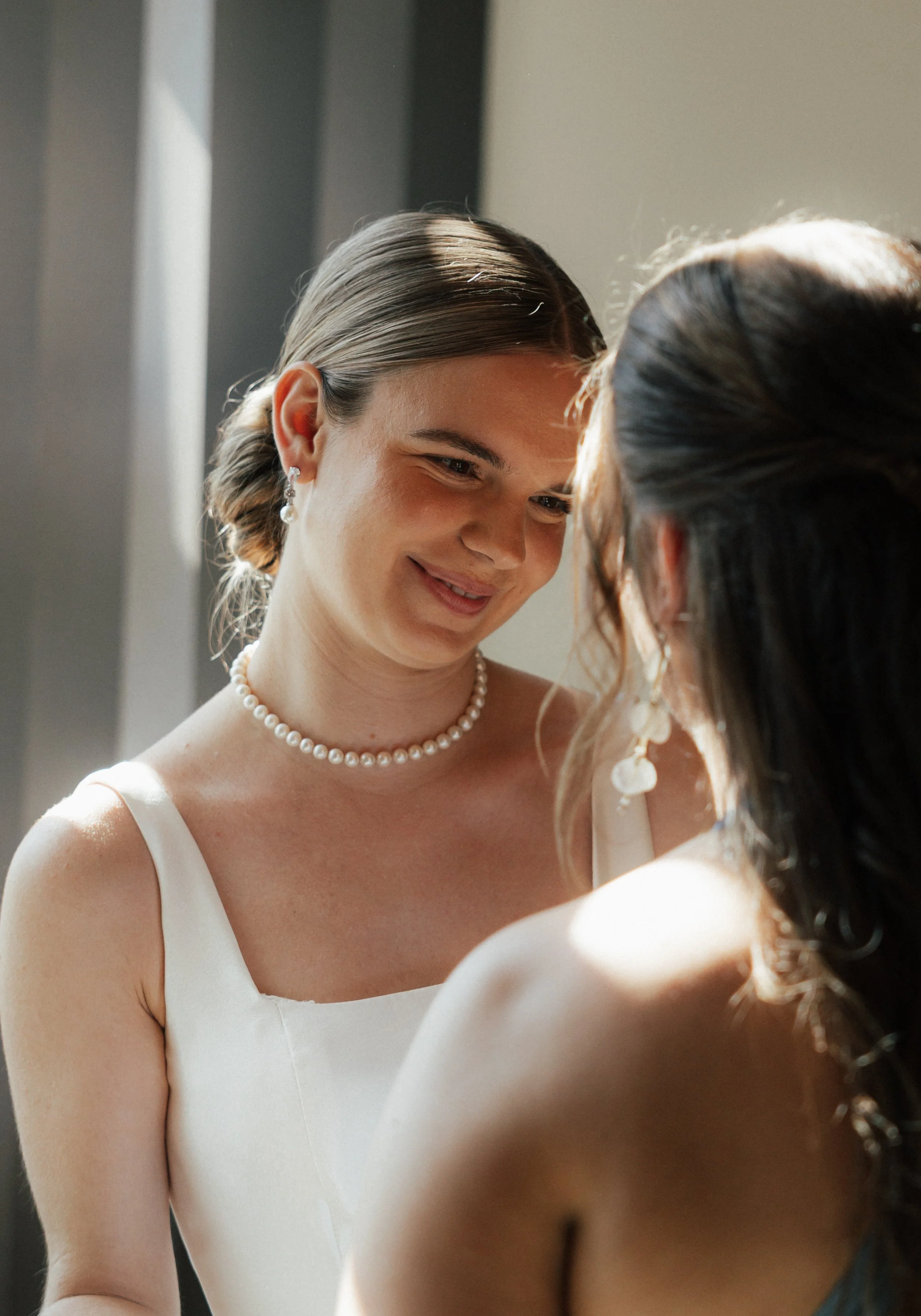 Two women in formal attire sharing a moment indoors, with one woman smiling softly at the other.