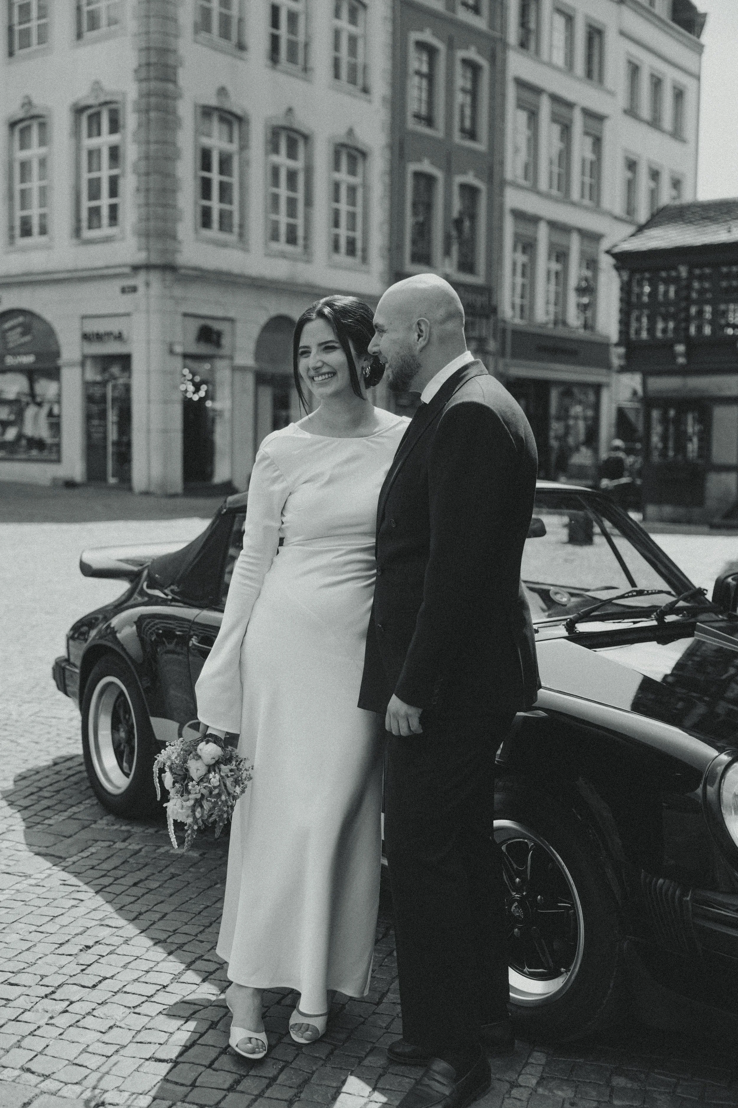 A bride and groom stand beside a vintage car on a city street, smiling and looking at each other. The bride holds a bouquet and wears a long white dress with bell sleeves, while the groom wears a dark suit. Buildings and storefronts are visible in the background.