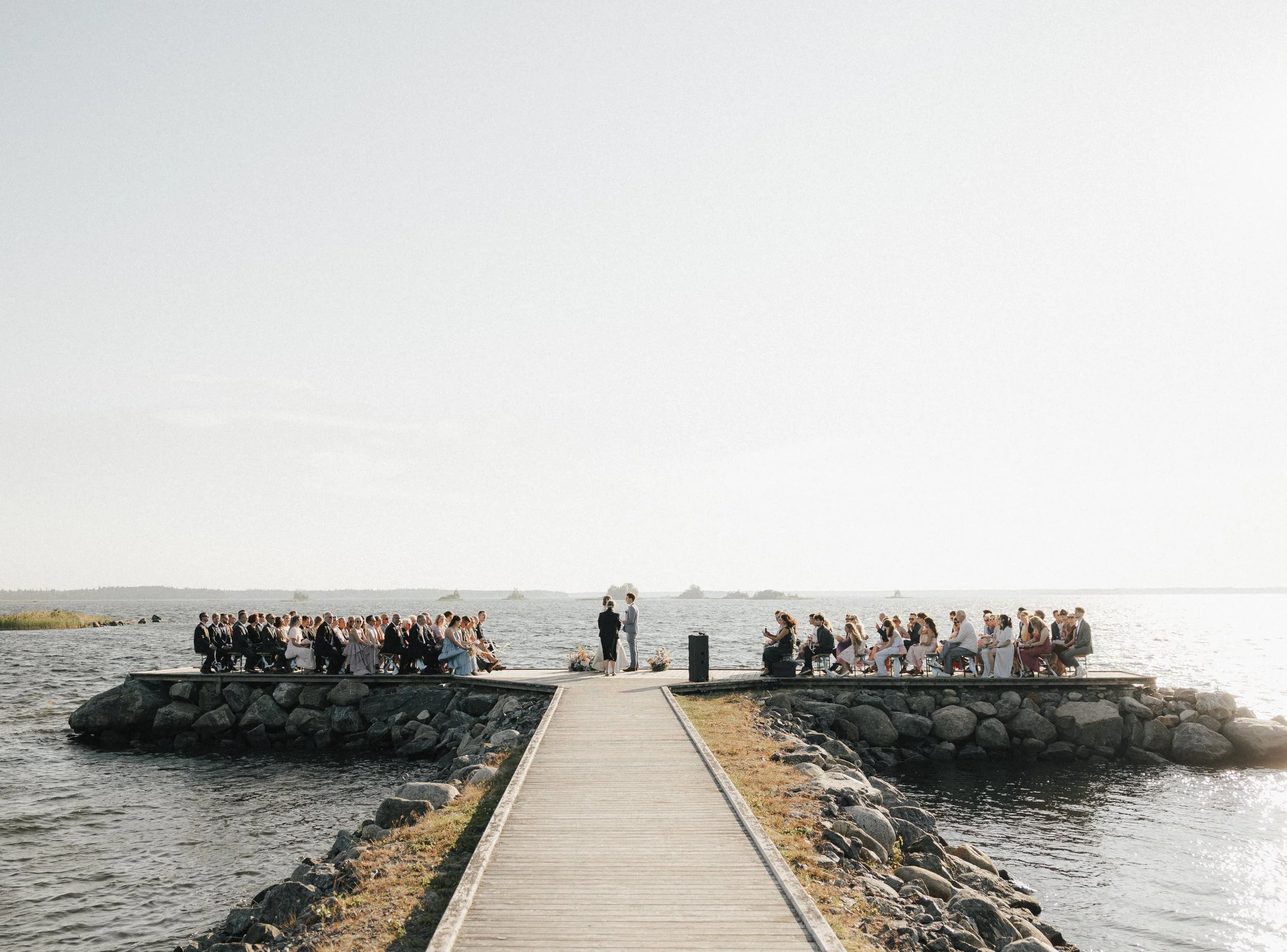 An outdoor wedding ceremony on a rocky pier over water, with attendees seated on both sides and a couple standing at the altar, with a clear sky and distant ships visible.