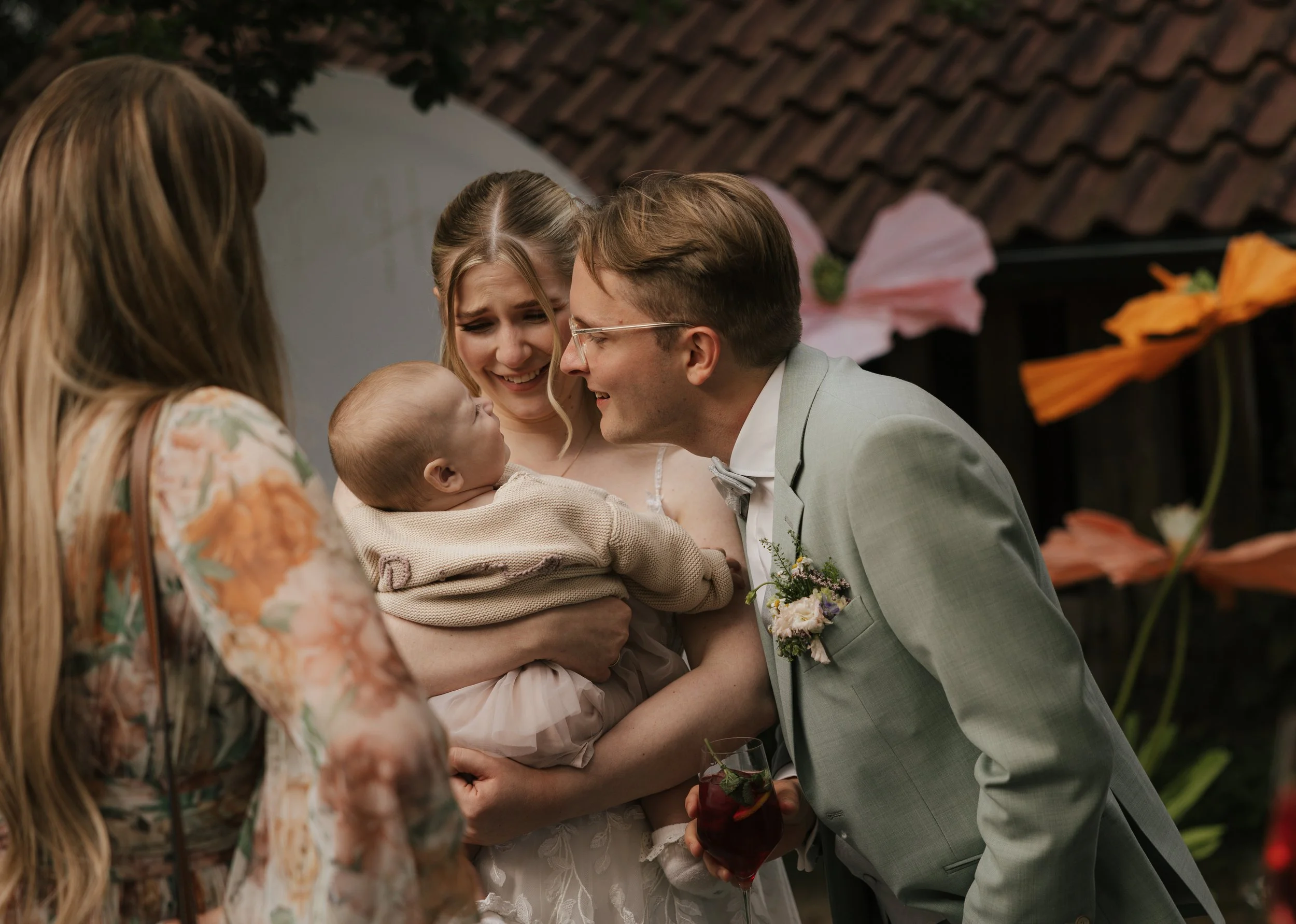 A group of four people at a wedding: a woman holding a baby, a man leaning in towards the baby, and a woman with long hair. The man is wearing a light gray suit with a boutonniere, and the women are in floral dresses. The background shows a building with a tiled roof and some large flowers.