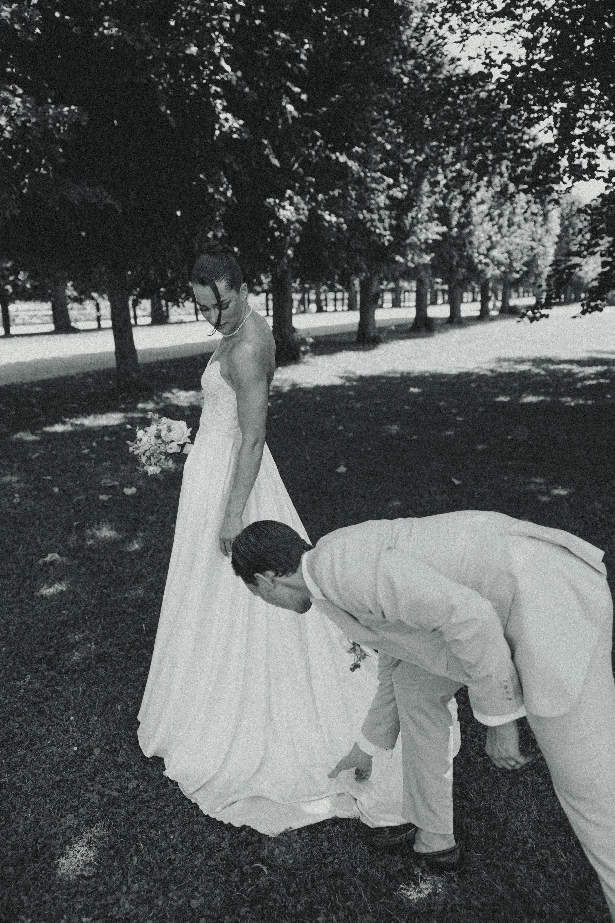 A black and white photo of a bride in a wedding dress with a groom in a suit outdoors under trees. The groom is bent down, pointing at the ground, while the bride holds a bouquet and looks down.