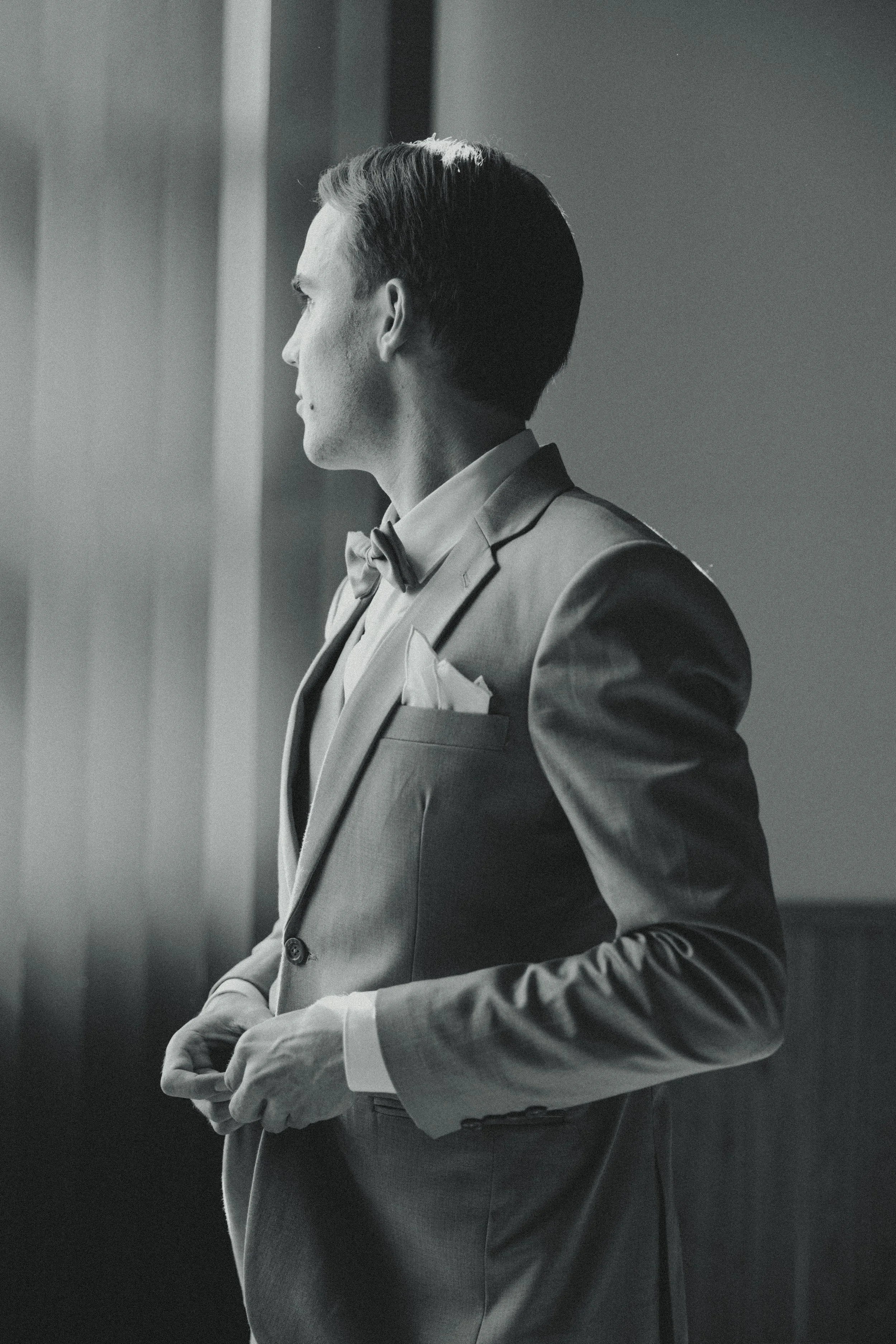 A black and white photo of a man in a tuxedo with a bow tie, standing and looking out a window.