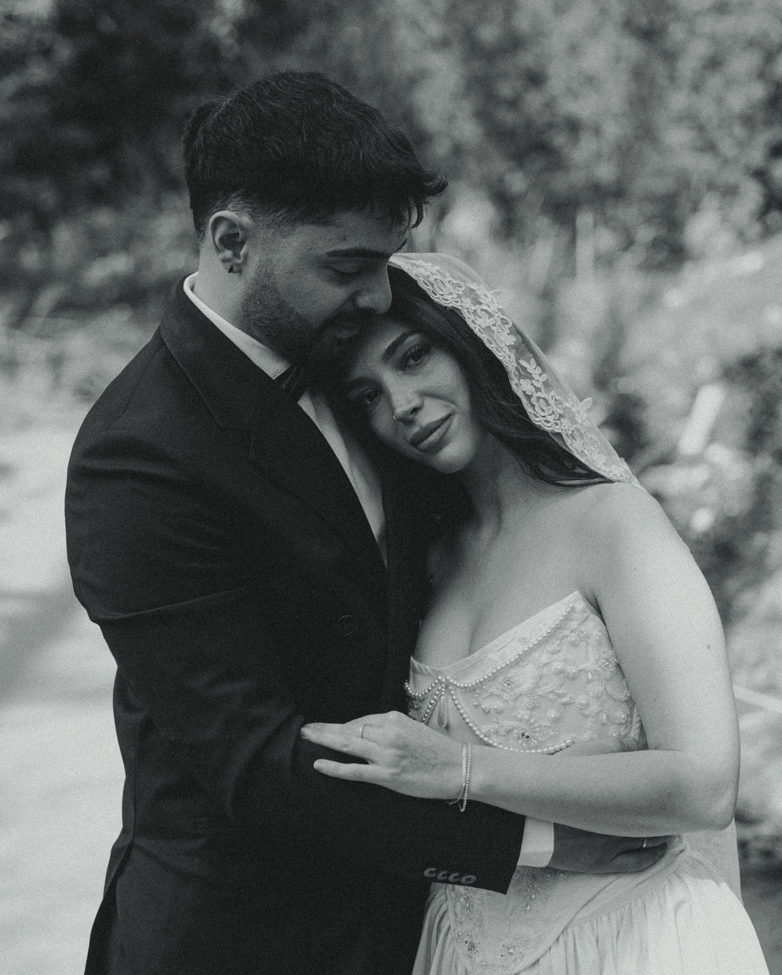 Black and white photo of a bride and groom embracing outdoors, with the bride wearing a strapless wedding dress and lace veil, and the groom in a suit with a bow tie.
