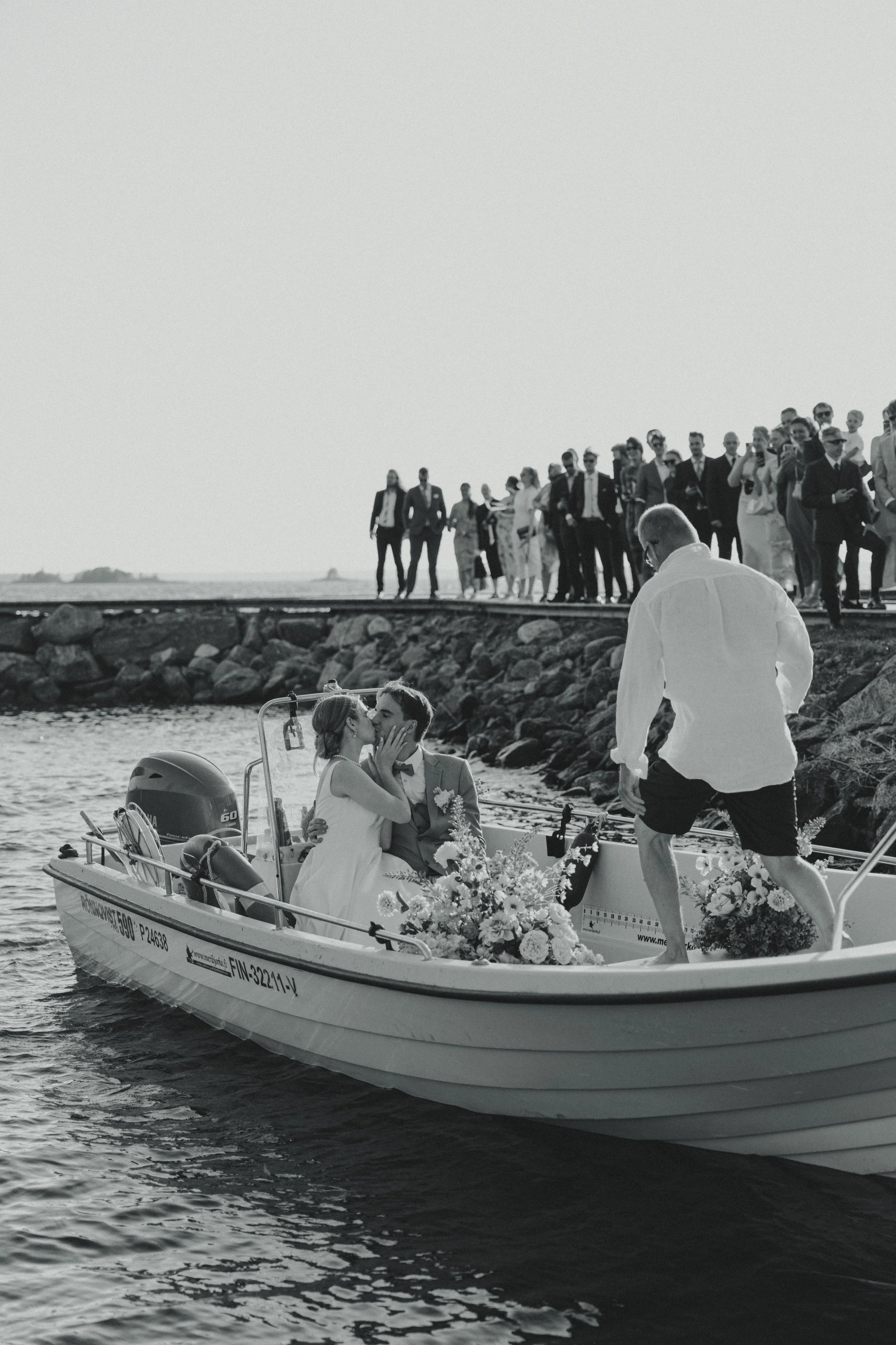 A couple in wedding attire on a boat, sharing a kiss, surrounded by flowers, with guests on the dock watching in the background. The scene is outdoors, near a body of water.
