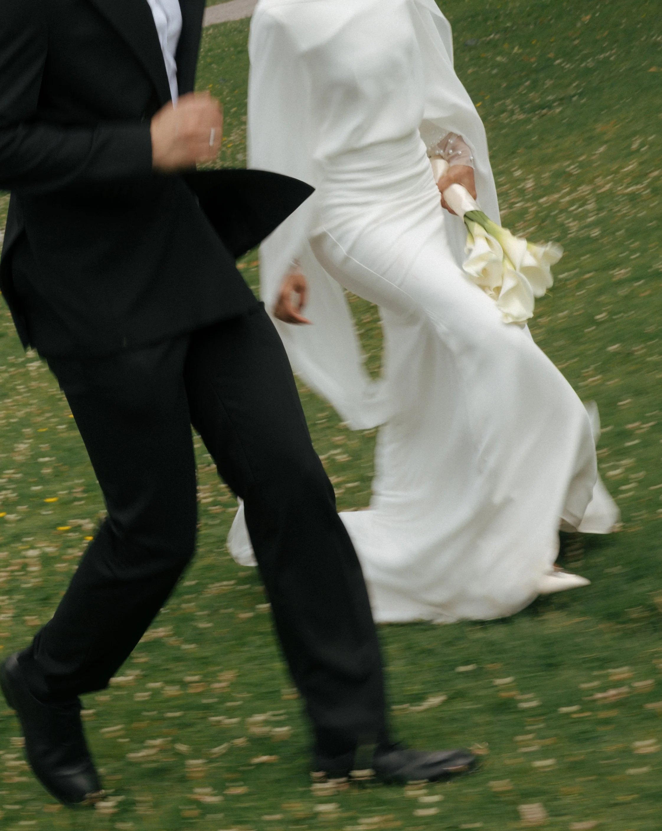 Close-up of a bride in a white wedding dress holding a bouquet of white calla lilies and a groom in a black suit walking on grass.