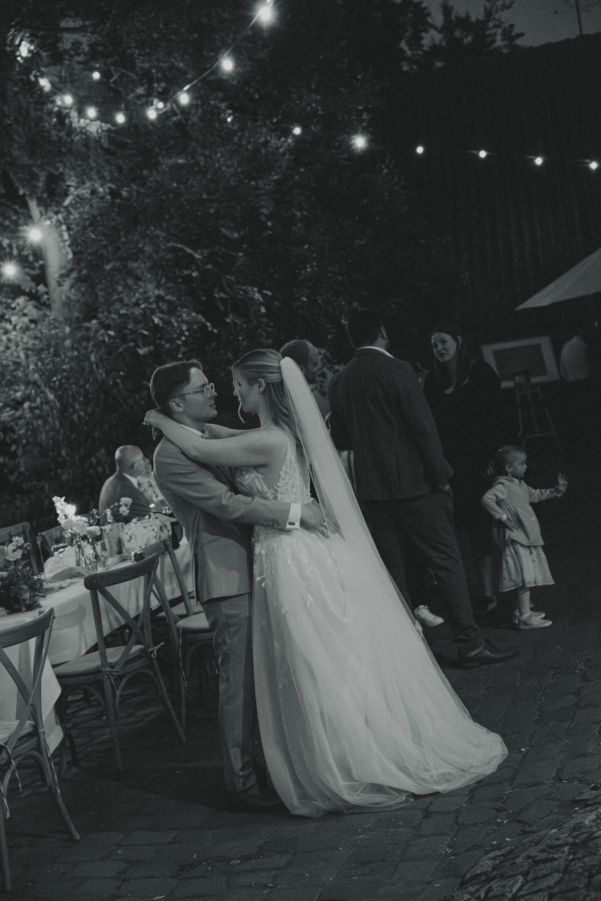 A black and white photo of a bride and groom dancing at an outdoor wedding reception at night, with string lights overhead and guests in the background.