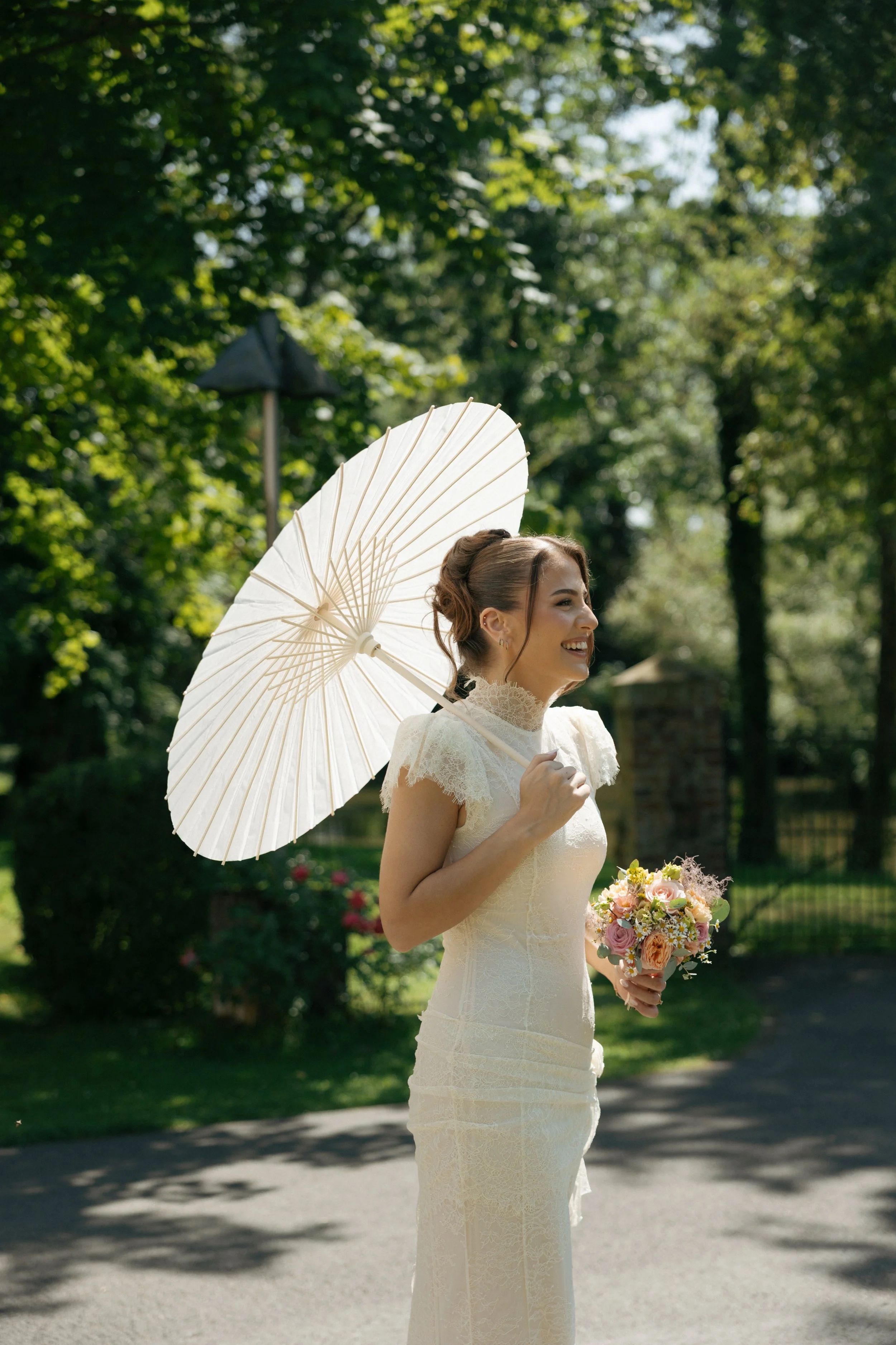 A smiling woman in a wedding dress holding a bouquet of flowers and an open white parasol outdoors with trees in the background.