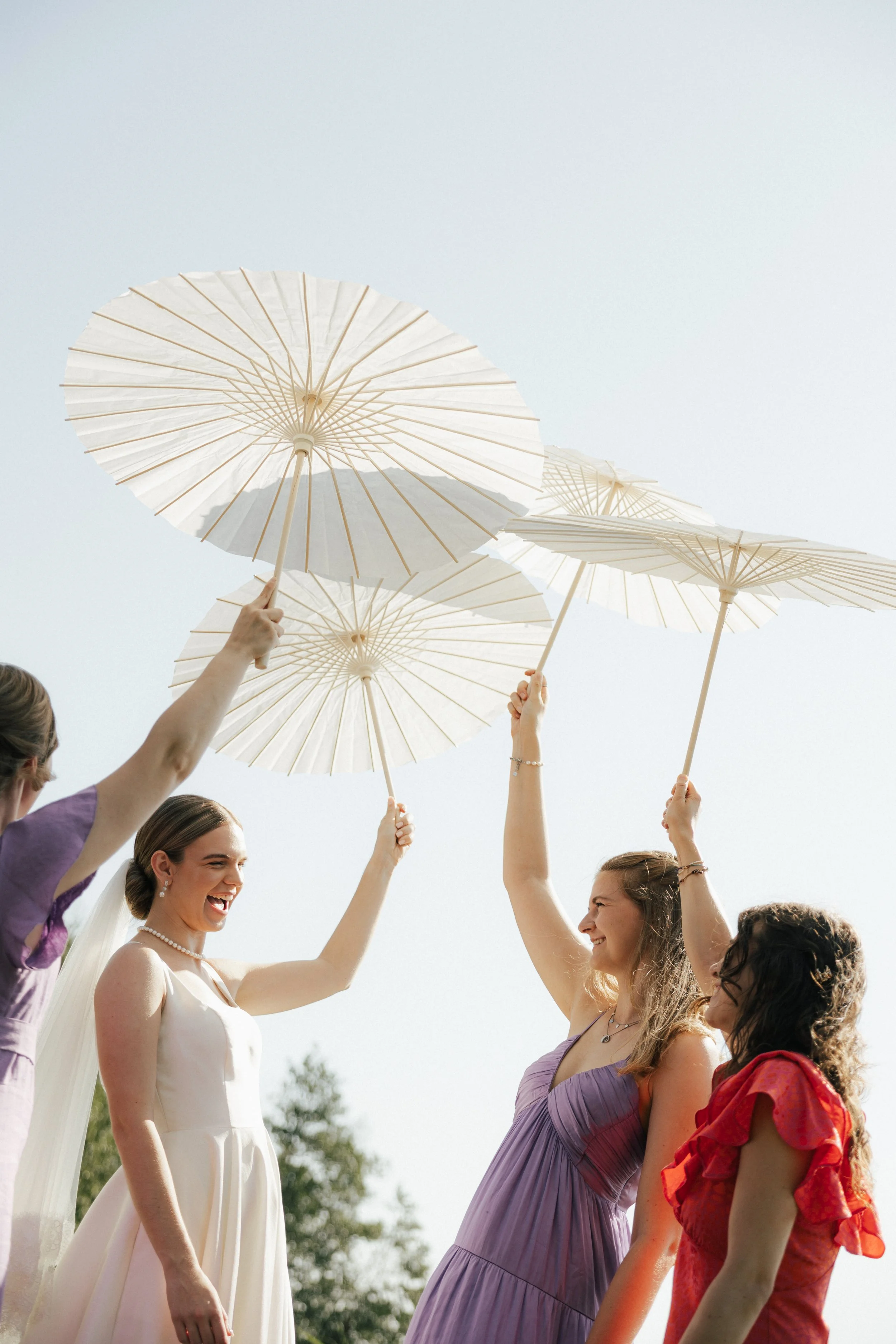 Four women smiling and holding white paper parasols up in the air, outdoors on a sunny day.