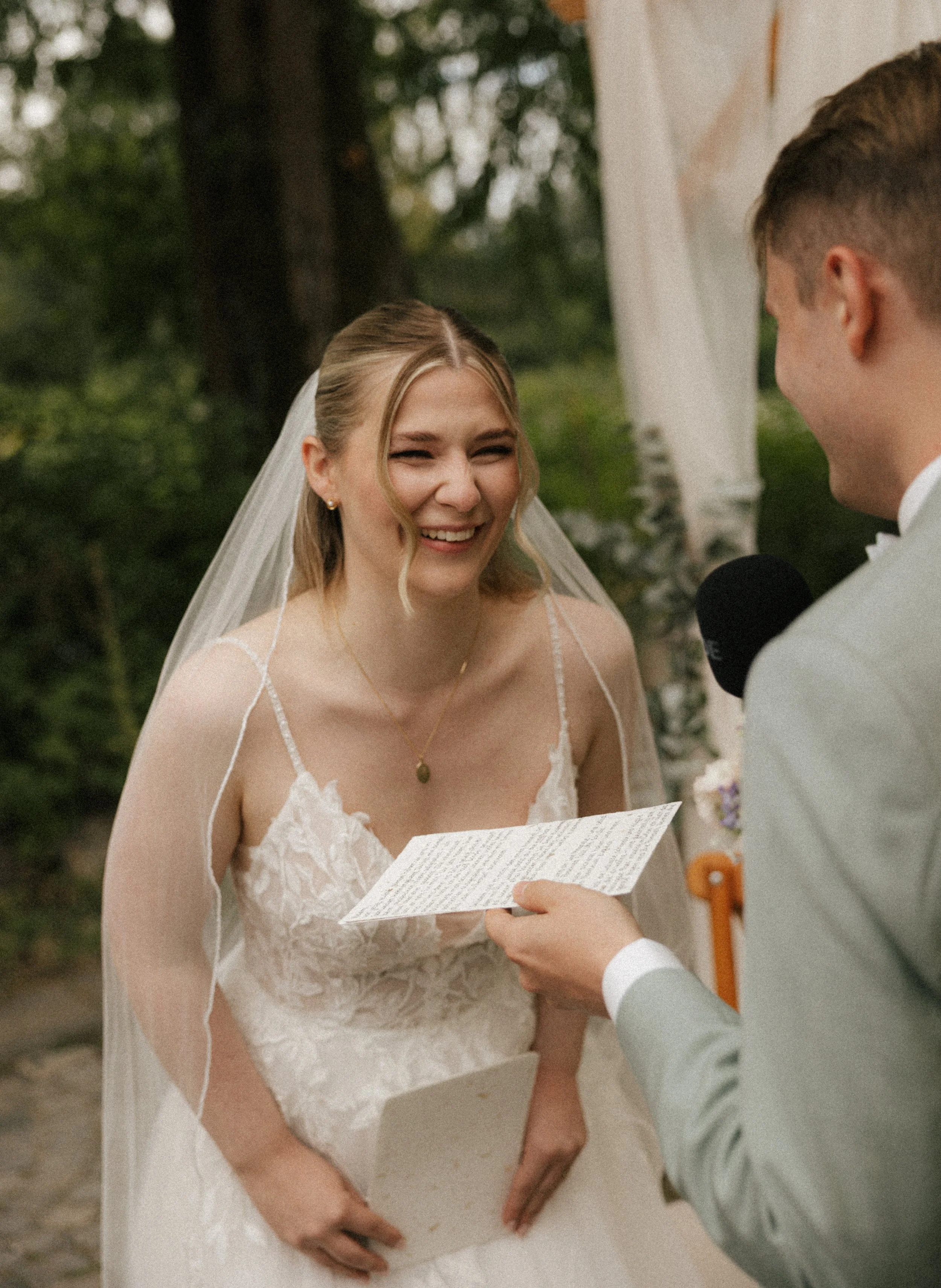 A bride smiling during her wedding ceremony, standing outdoors, wearing a lace wedding dress and veil, holding a paper, with a groom reading vows.