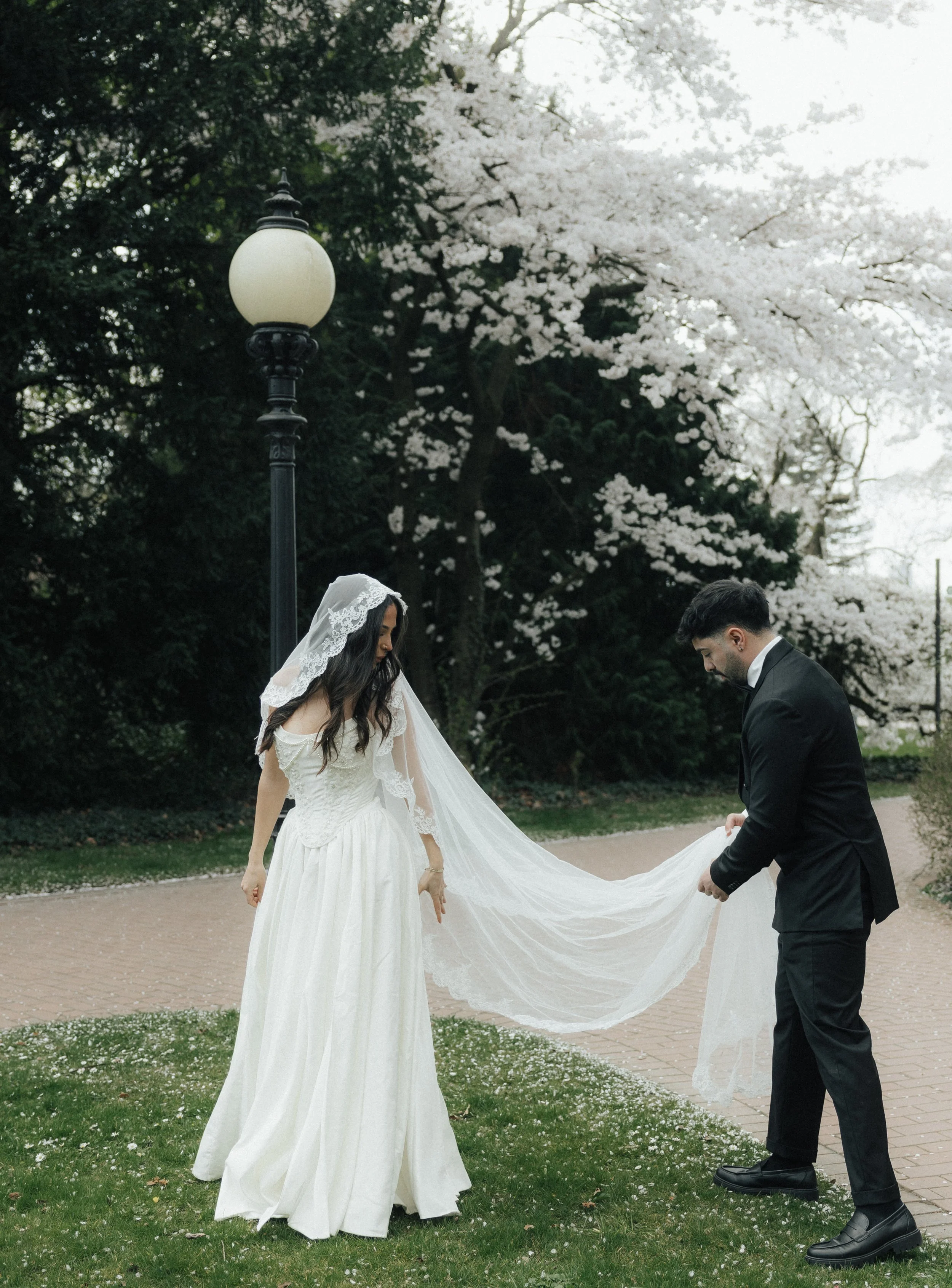 A bride and groom in a garden with blooming trees, with the groom holding the bride's train and both wearing formal wedding attire.