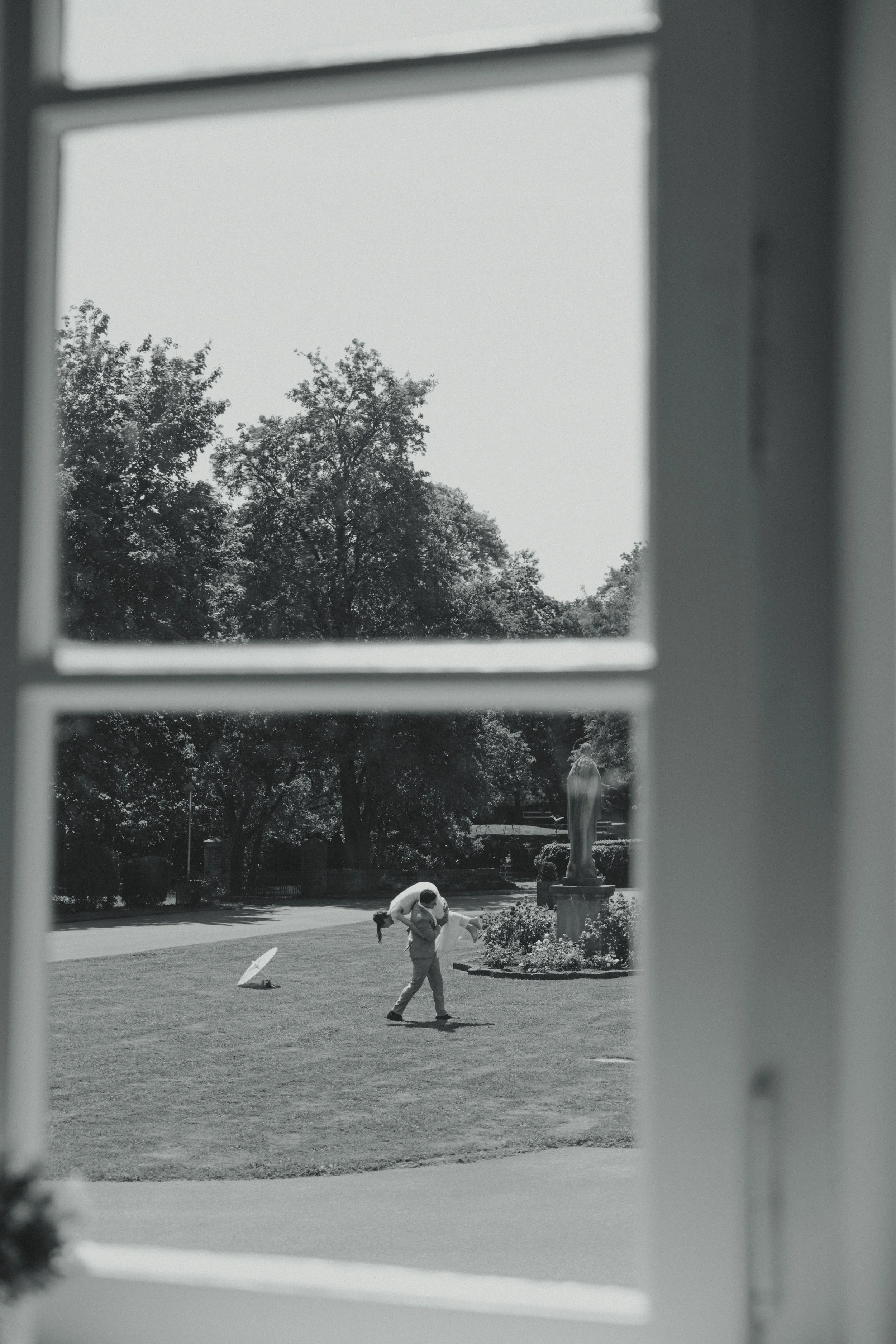 View through a window showing a person carrying another person on their back in an outdoor park with trees and a statue in the background.