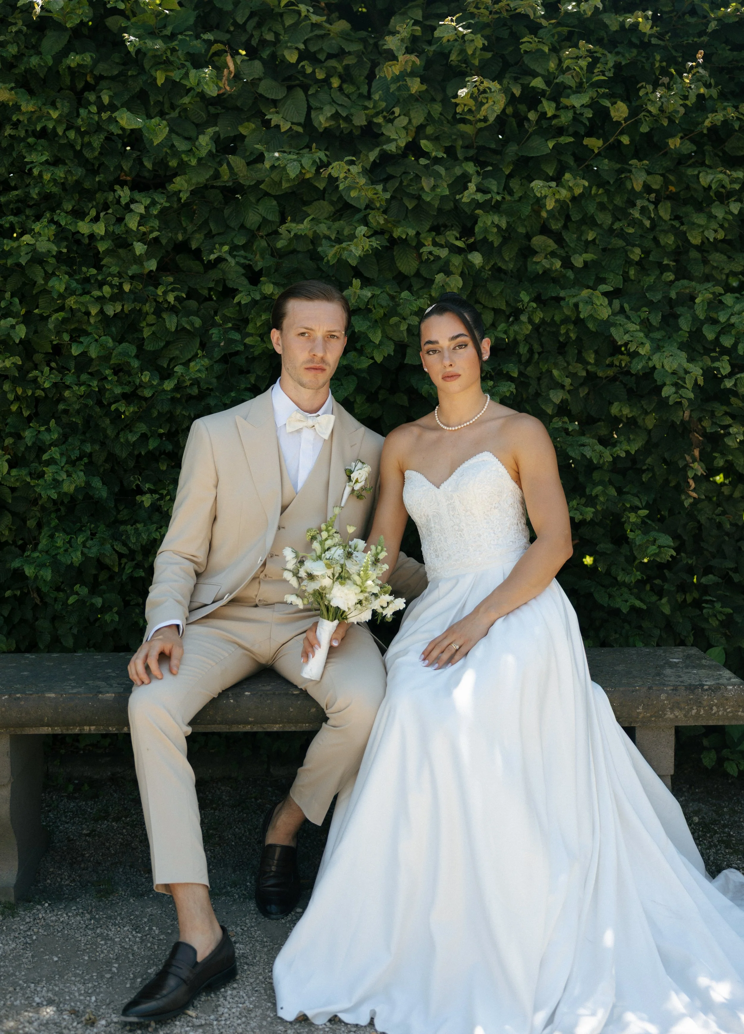 A bride and groom sitting on a stone bench outdoors, the groom in a beige suit and the bride in a strapless white wedding dress, holding a bouquet of white flowers against a green leafy background.