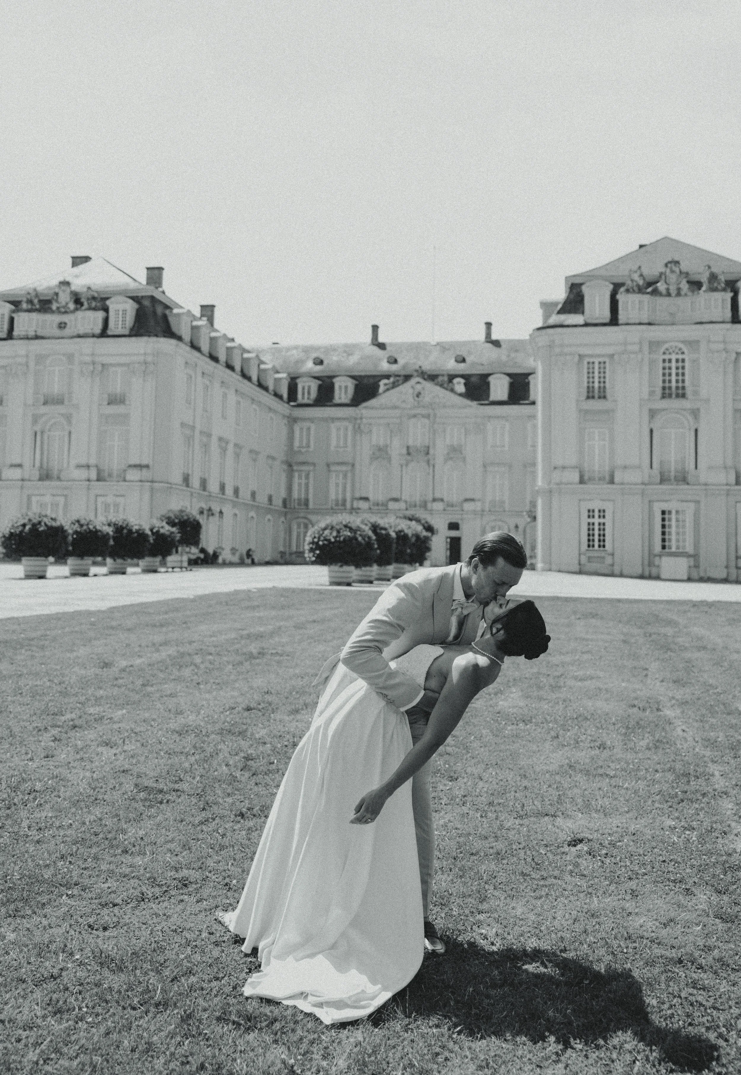 A black-and-white photo of a couple dancing in front of a large, historic building with ornate architecture and well-kept gardens.