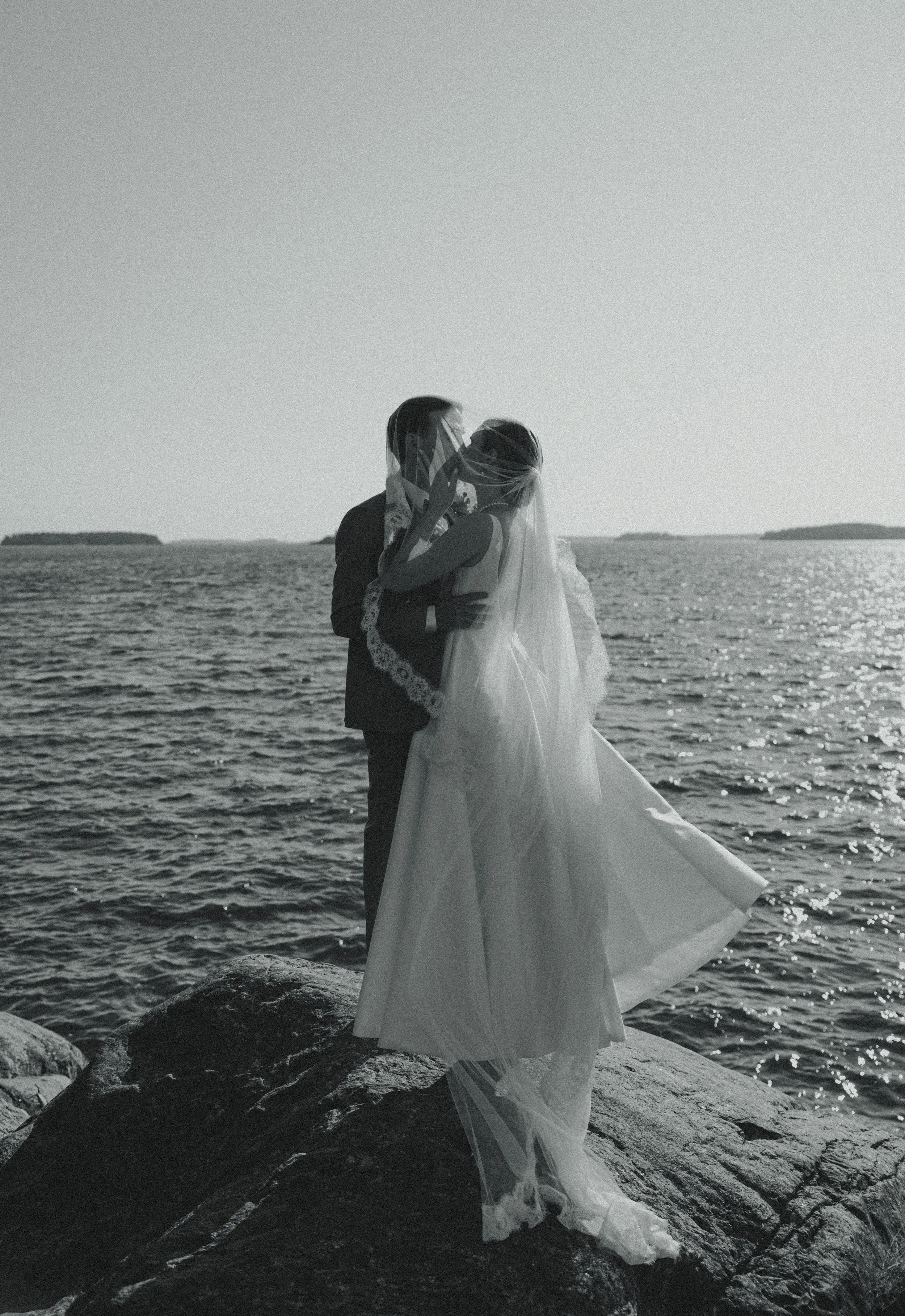 A couple in wedding attire embracing by the water, with the bride wearing a veil and flowing dress, and the groom in a suit, during sunset or sunrise.