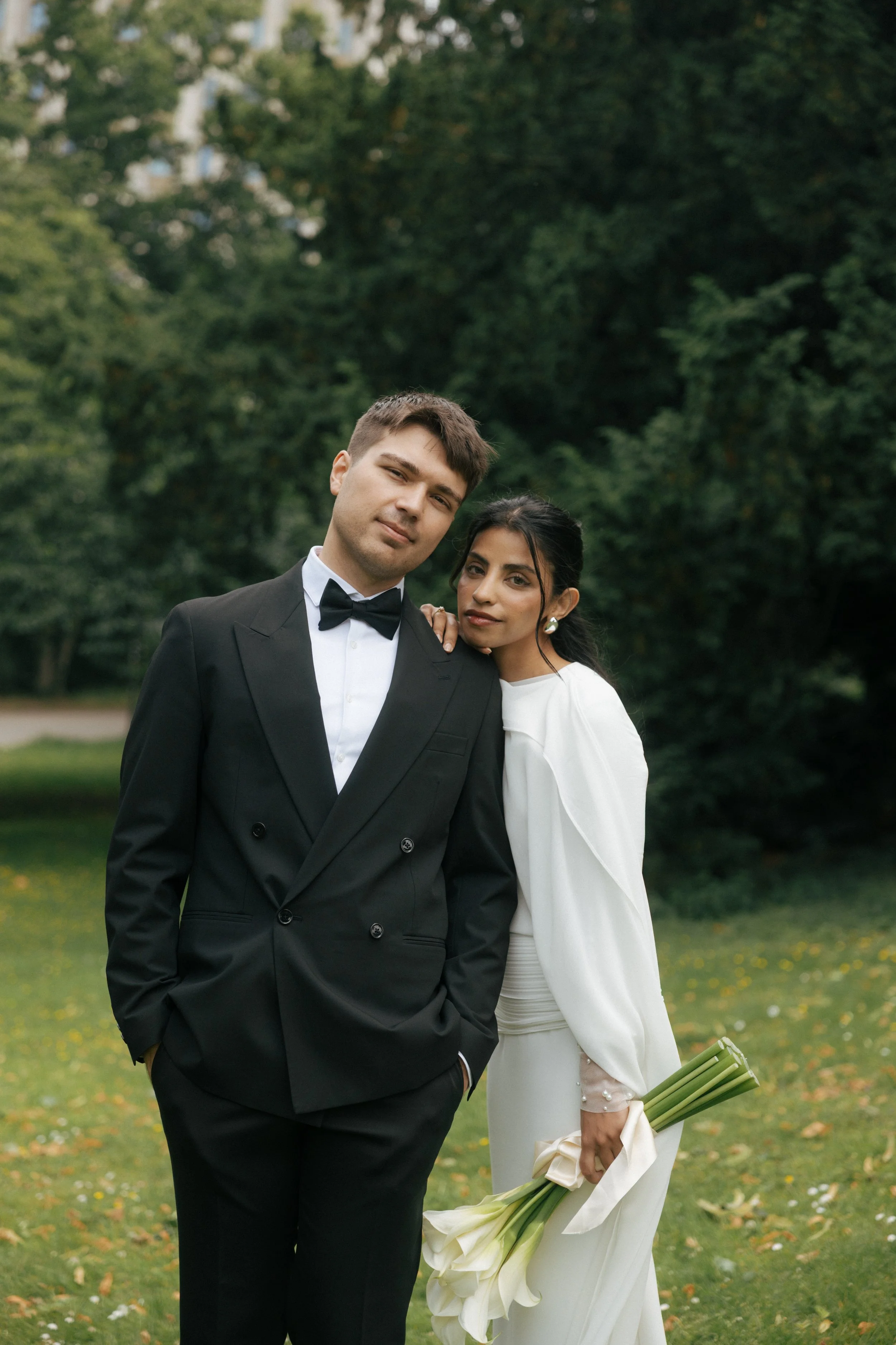 A couple dressed in formal wedding attire standing outdoors on a grassy area, with the man in a black tuxedo and the woman in a white dress holding a bouquet of white calla lilies.