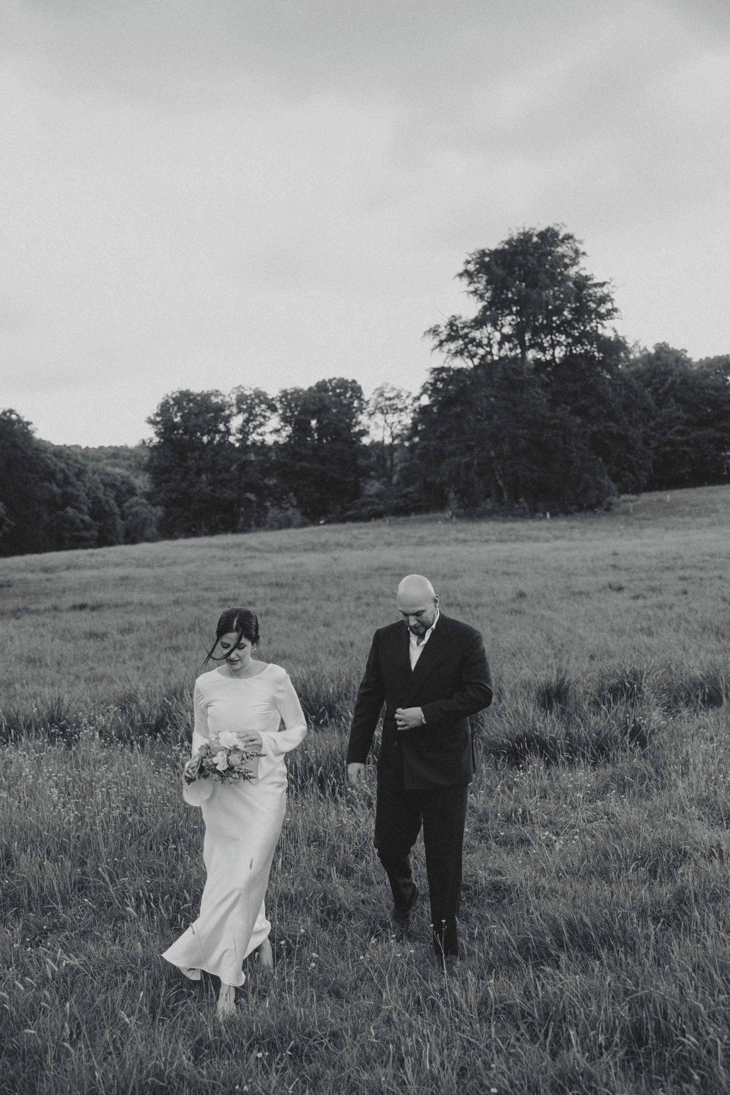 A black and white photo of a bride in a long white dress holding a bouquet and a groom in a dark suit walking through a grassy field with trees and cloudy sky in the background.