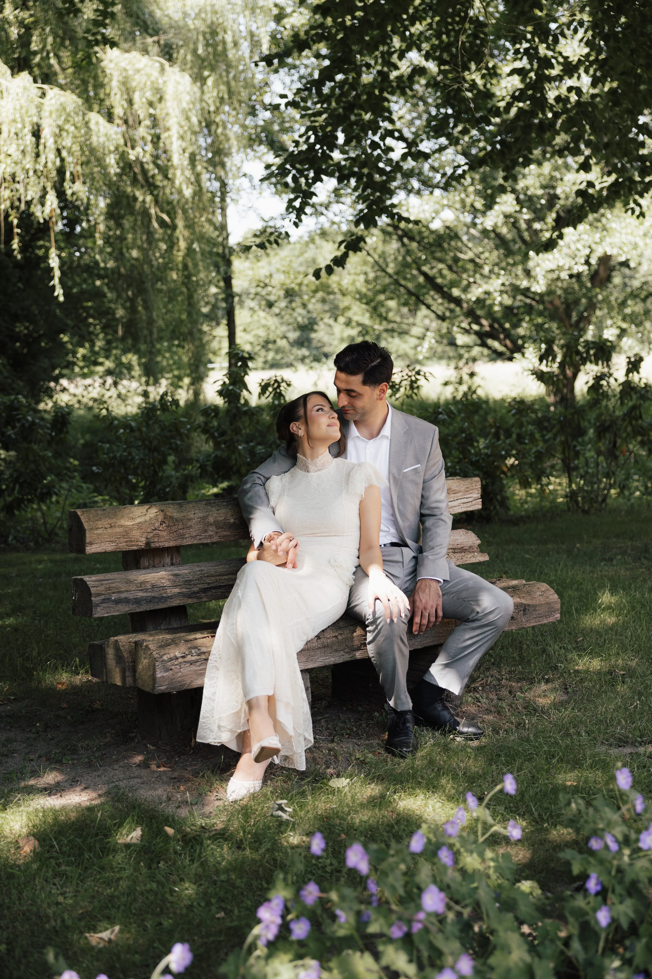 A couple dressed in wedding attire sitting on a wooden park bench, surrounded by lush greenery and small purple flowers, sharing an intimate moment.