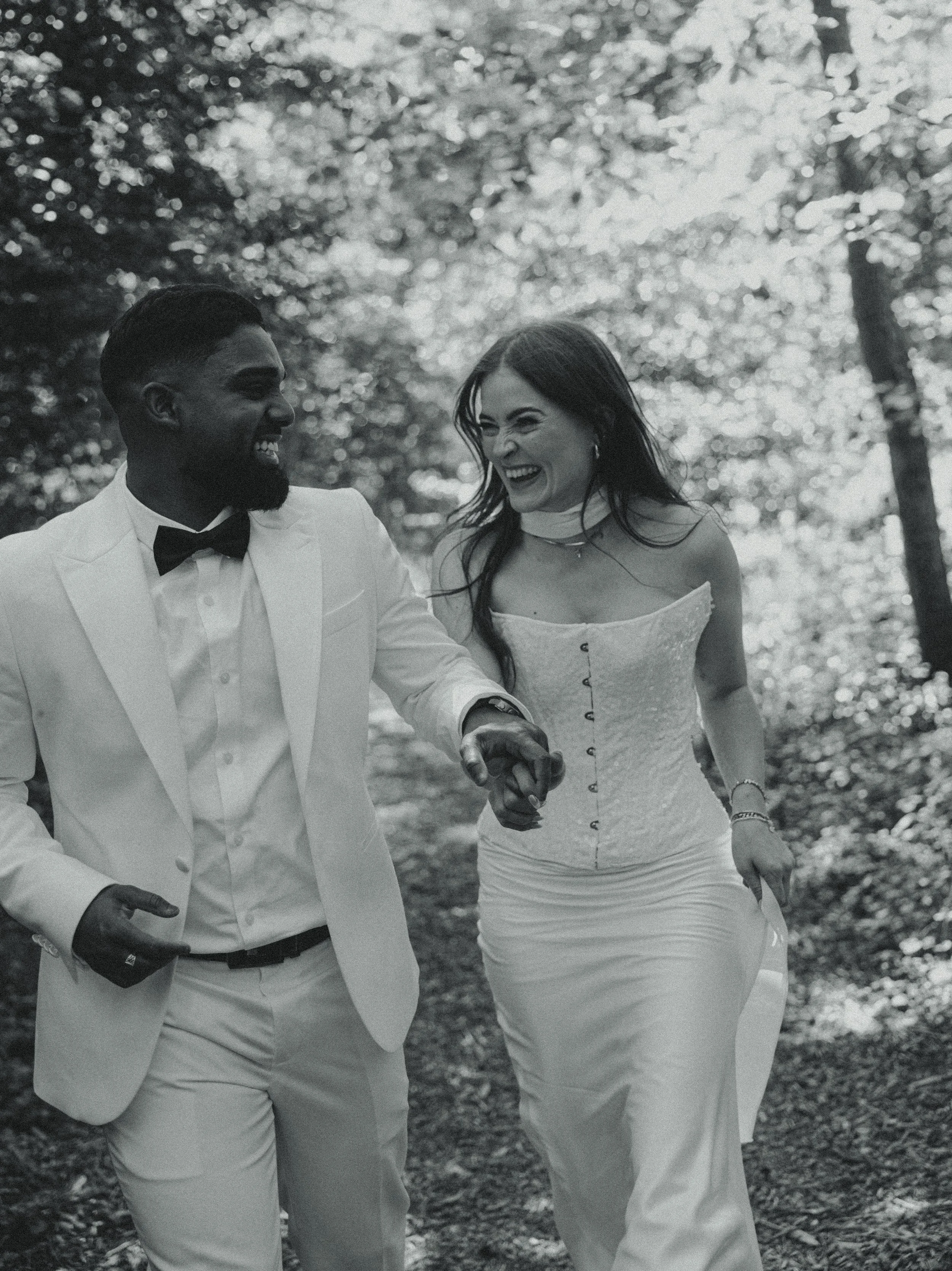 A smiling couple, the man in a white tuxedo with a bow tie and the woman in a strapless wedding dress, holding hands and walking through a wooded area.