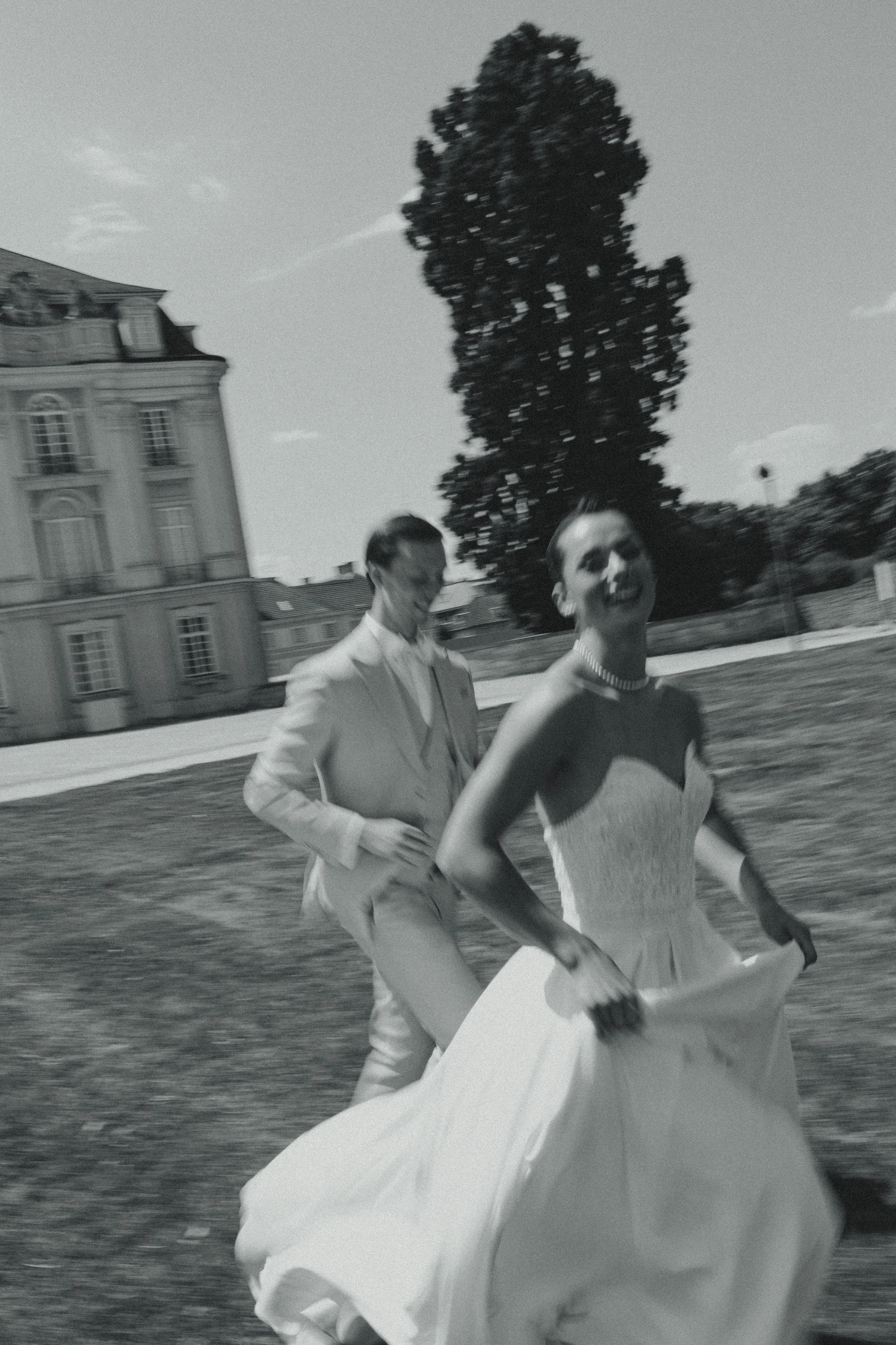 A black-and-white photo of a man and woman outdoors, with the man in a light suit and the woman in a strapless gown holding the skirt, smiling and appearing joyful. There is a large building and a tall tree in the background.
