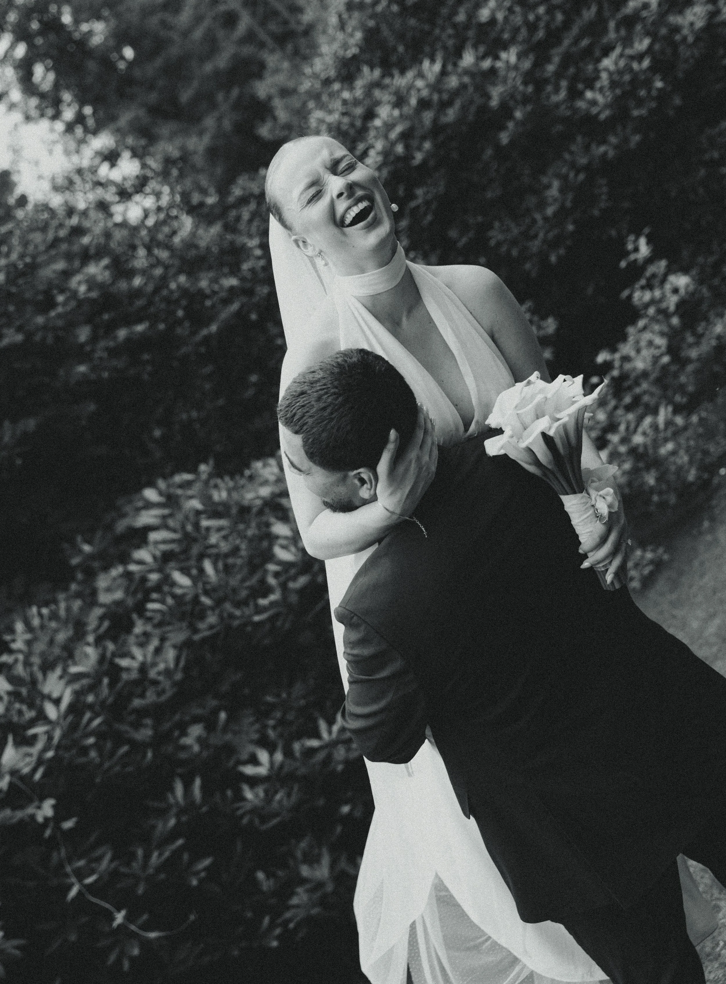A bride laughing joyfully while holding a bouquet, being kissed or embraced by her groom in an outdoor setting with trees and foliage in the background, captured in black and white.