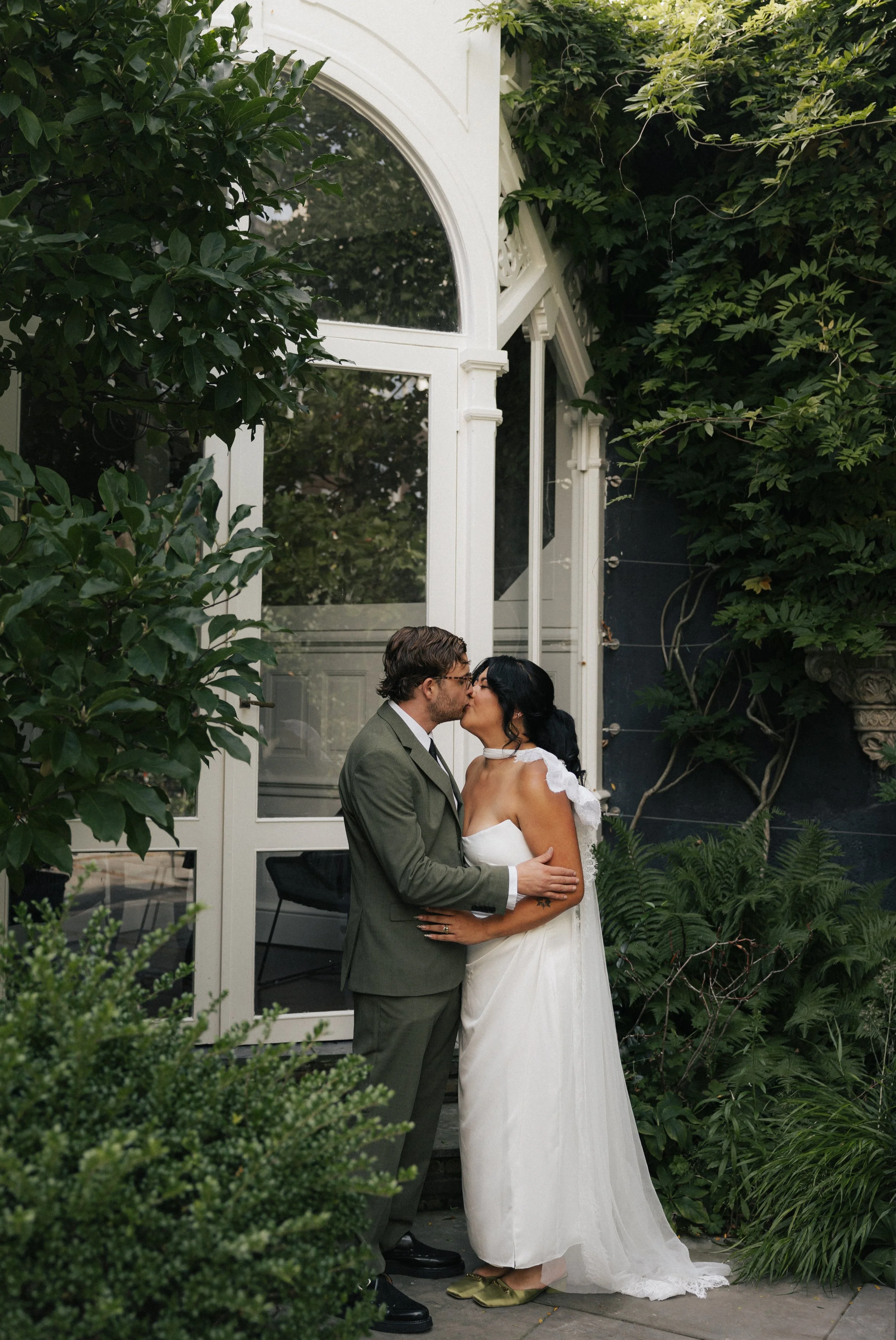 A couple in wedding attire sharing a kiss outdoors, surrounded by lush green plants and a glass structure with white framing.