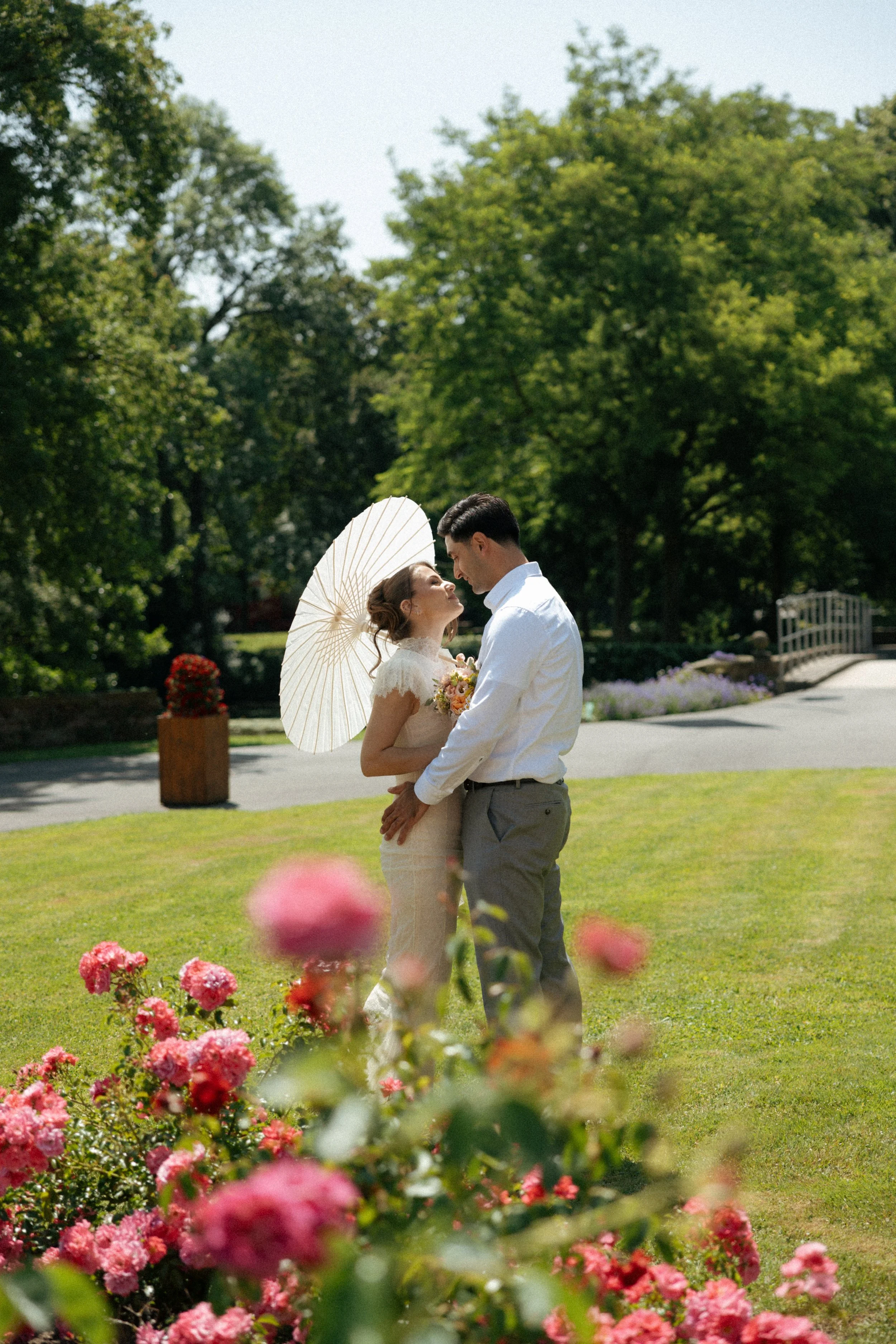 A bride and groom standing close together on a sunny day in a garden, with pink flowers in the foreground. The bride holds a white parasol, and they are gazing at each other affectionately.