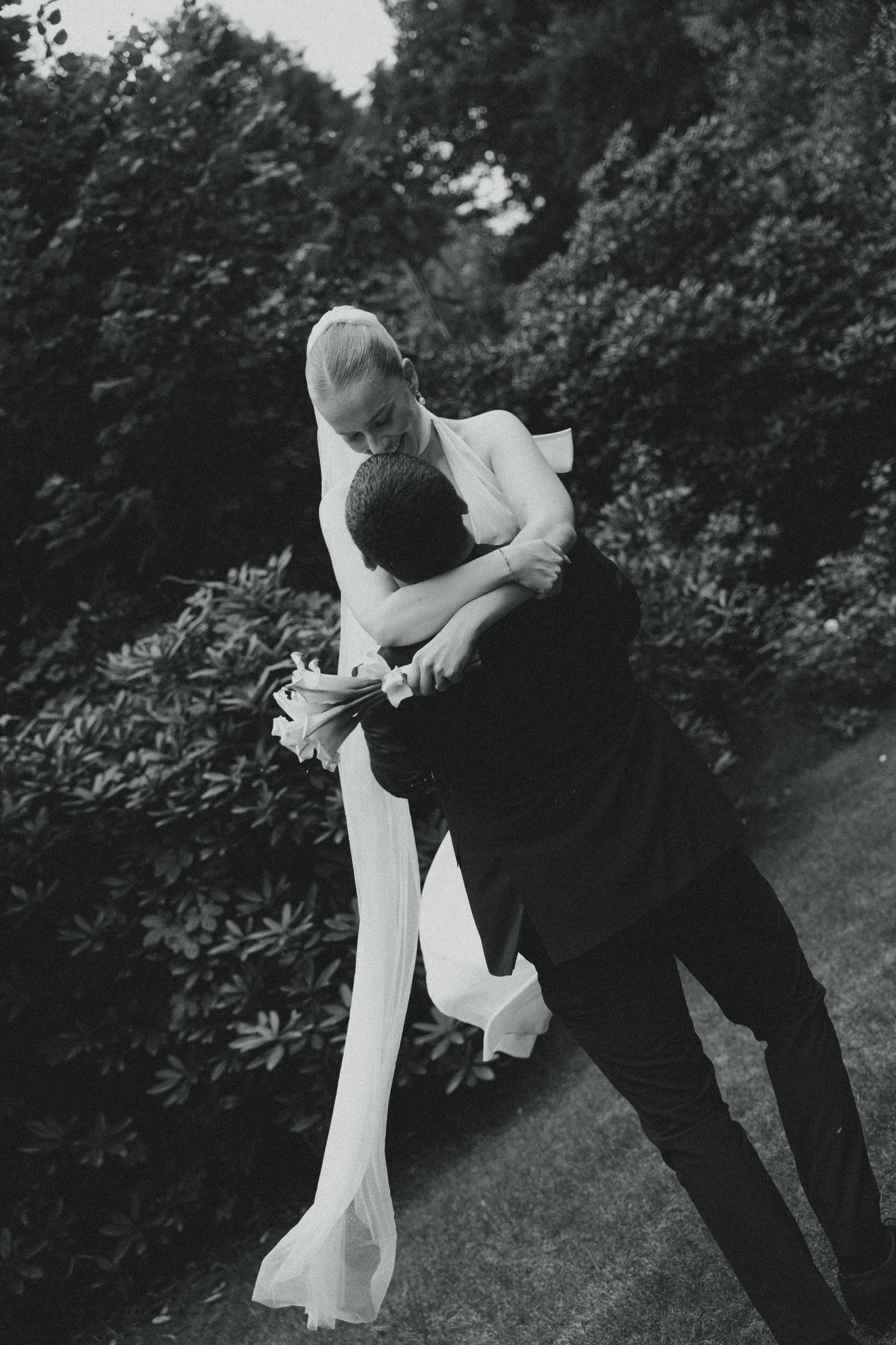 A bride in a wedding dress embracing a groom in a suit outdoors, holding a bouquet of flowers.