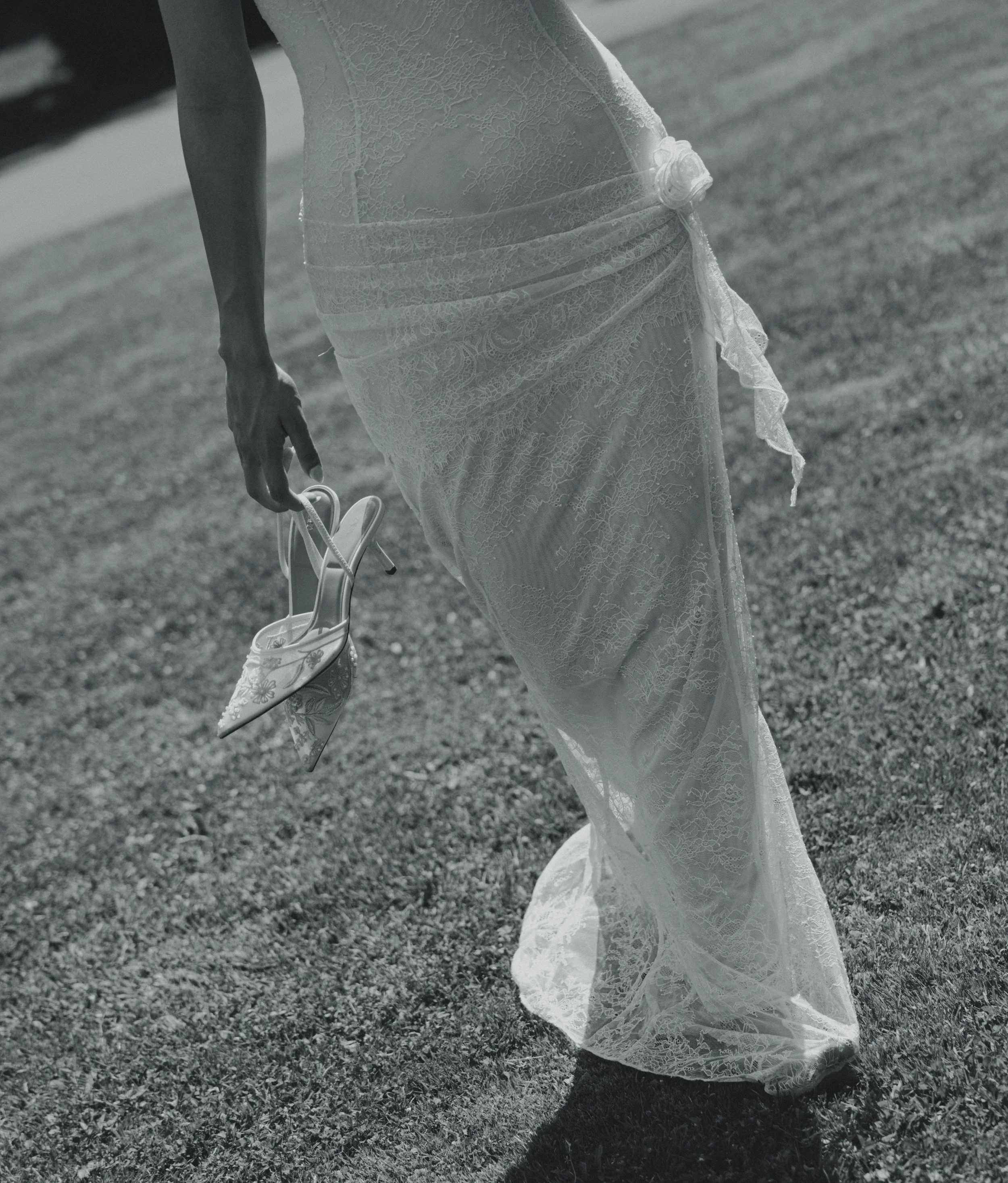 A woman in a lace dress holding a pair of striped high-heeled shoes on a grassy surface.